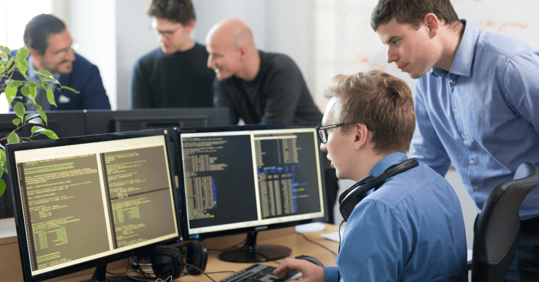 Engineering team collaborating at a computer with an overlay illustrating standardized AI workflows, shared prompt libraries, reusable AI‑assisted workflows, testing, validation, debugging, and deployment of AI playbooks across software development teams.