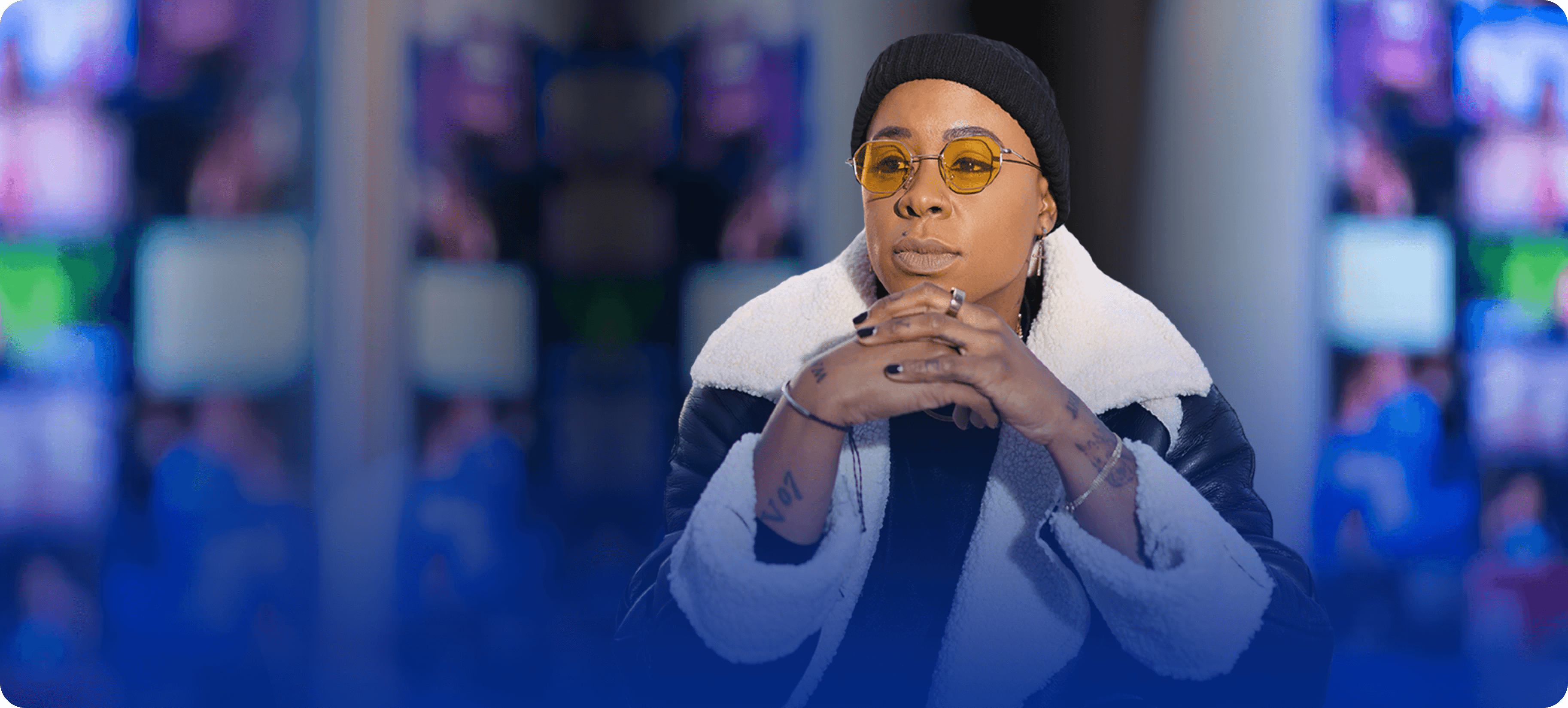 Ashleigh Warren, a person sits thoughtfully in front of multiple colorful screens, dressed in a stylish jacket.