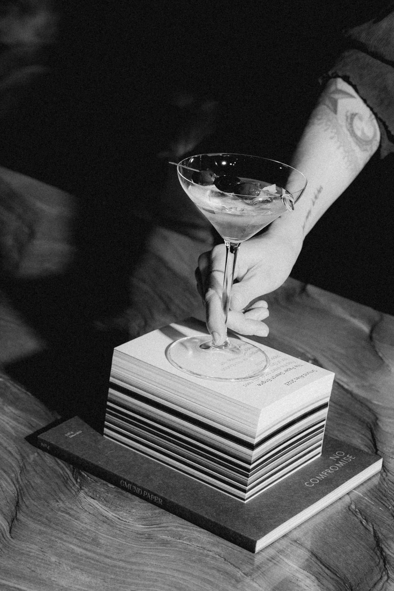 Schwarz-Weiß-Fotografie einer Hand, die ein Martini-Glas auf einem Stapel Bücher hält, mit dem Schriftzug 'MARTINI'