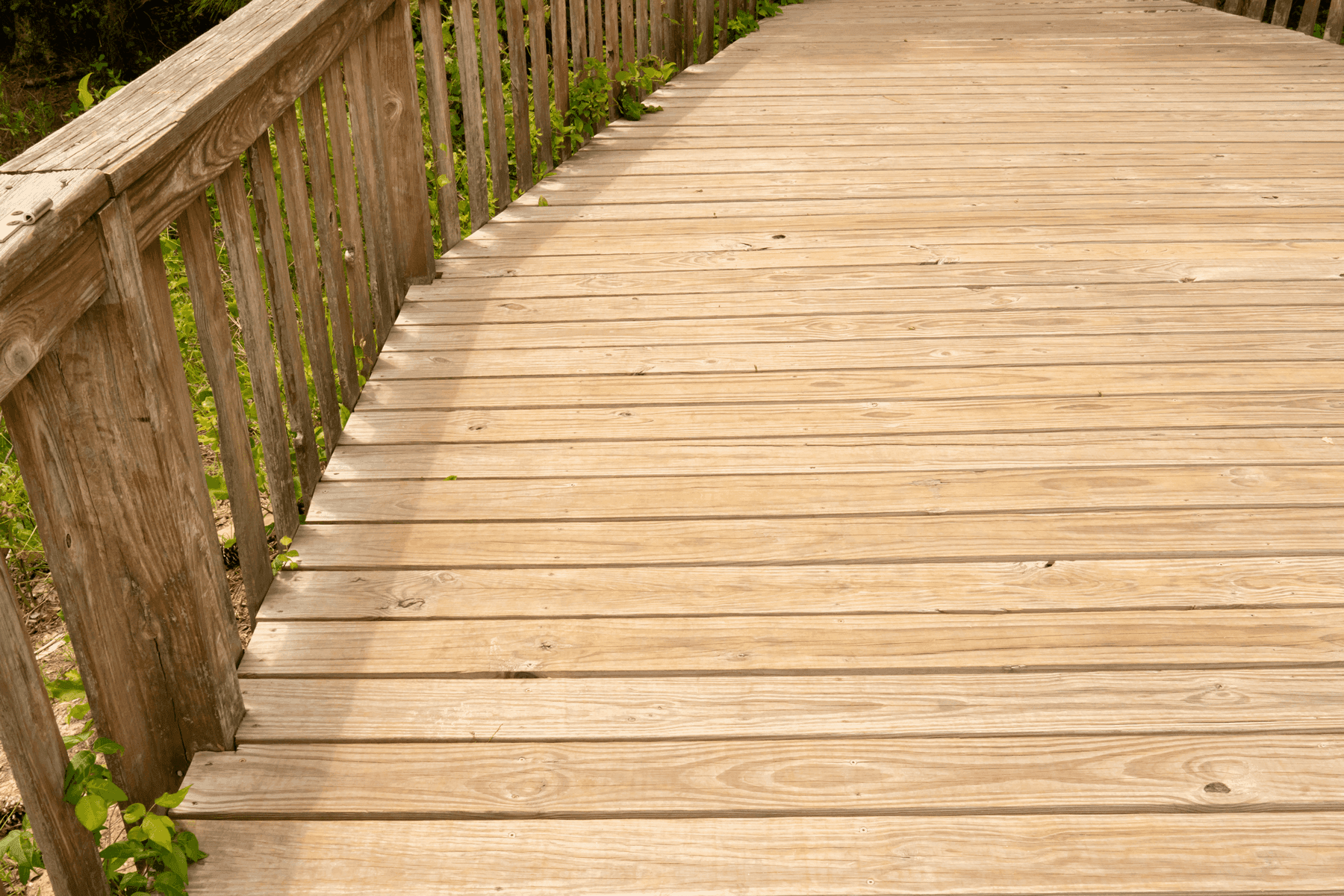 A weathered wooden boardwalk-style deck with natural wood tones and railings on one side. The deck is surrounded by greenery, giving it a rustic, natural feel.