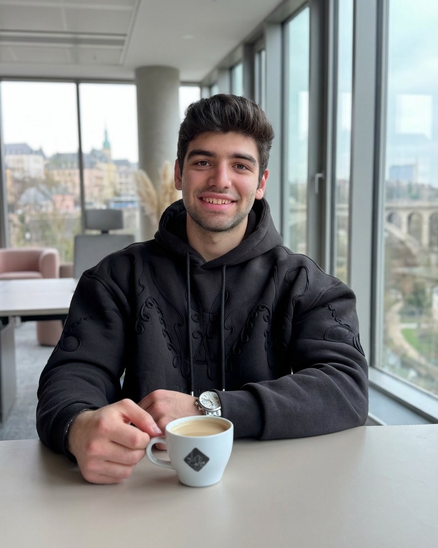 A young man in a red blazer and navy turtleneck, with wavy brown hair, poses against a neutral gray background.