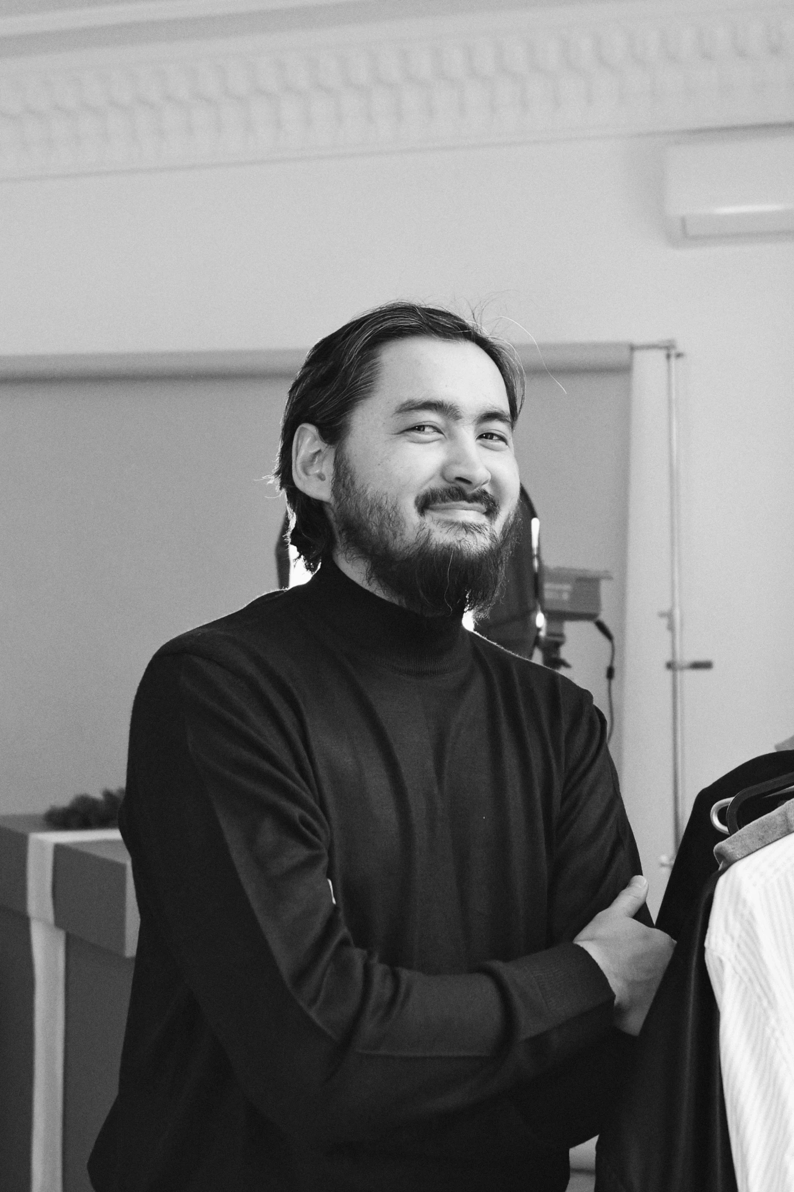 An architect standing at a desk in a modern studio, reviewing architectural models and documents. Shelves filled with books and materials line the background, with natural light coming through large windows.