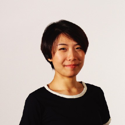 A young woman with short, dark hair styled in a pixie cut is captured against a plain off-white background. She is wearing a black top with a white trim around the neckline. Her gaze is directed towards the camera, and she has a slight smile. The lighting is even and highlights her features clearly.