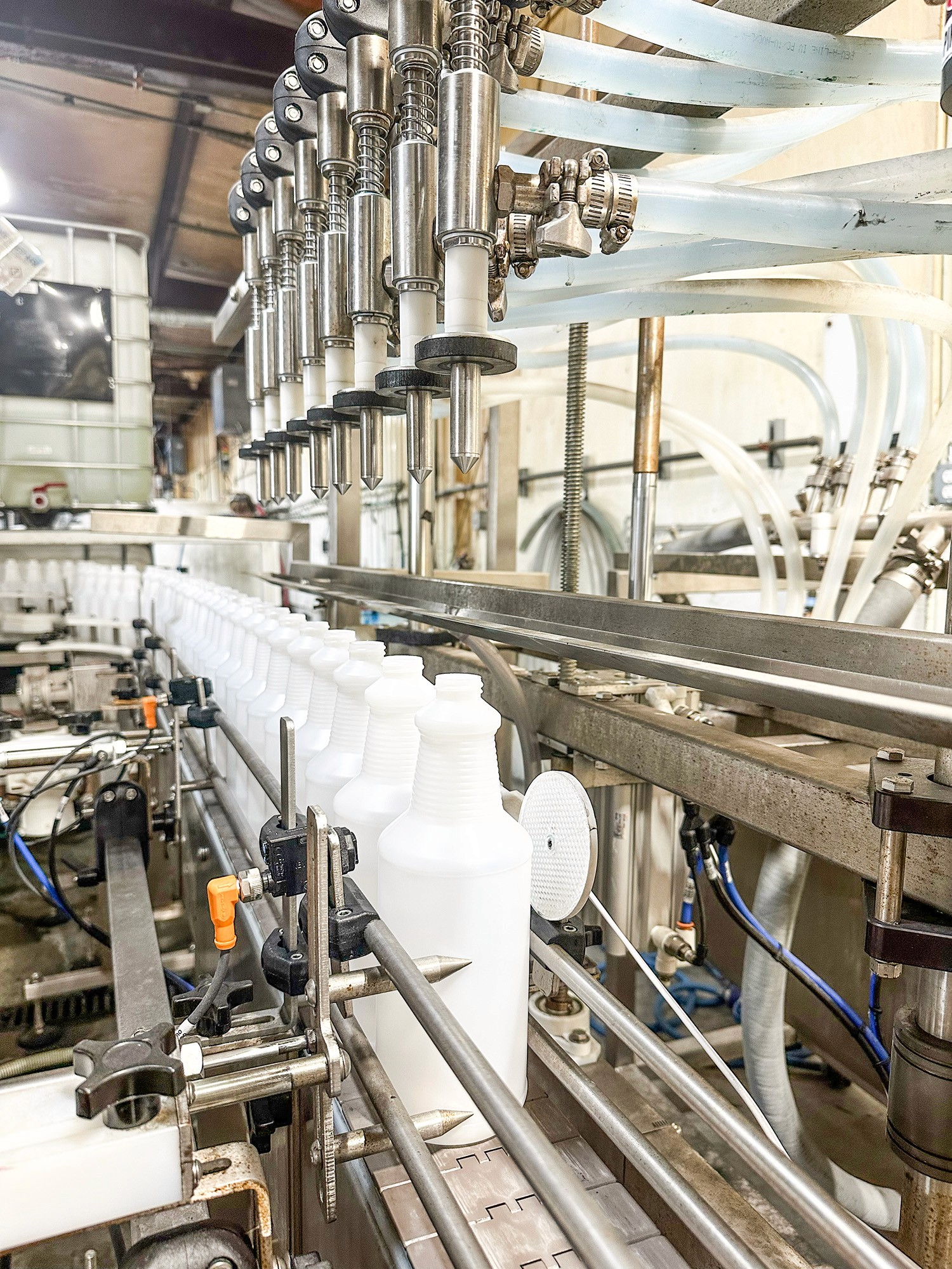 A detailed close-up view of an industrial bottling line in a factory setting. The image captures a row of white plastic bottles, resembling milk jugs, moving along a stainless steel conveyor belt. Above the bottles, a series of metallic filling nozzles, each connected to clear plastic tubing, are positioned to dispense liquid into the containers. The machinery is intricate, with visible springs, valves, and fittings, showcasing the precision of the automated filling process. The background includes additional industrial equipment and pipes, with a slightly blurred view of a white storage tank, emphasizing the clean, utilitarian environment of the production facility. The lighting is bright, highlighting the metallic sheen of the machinery and the smooth texture of the plastic bottles.