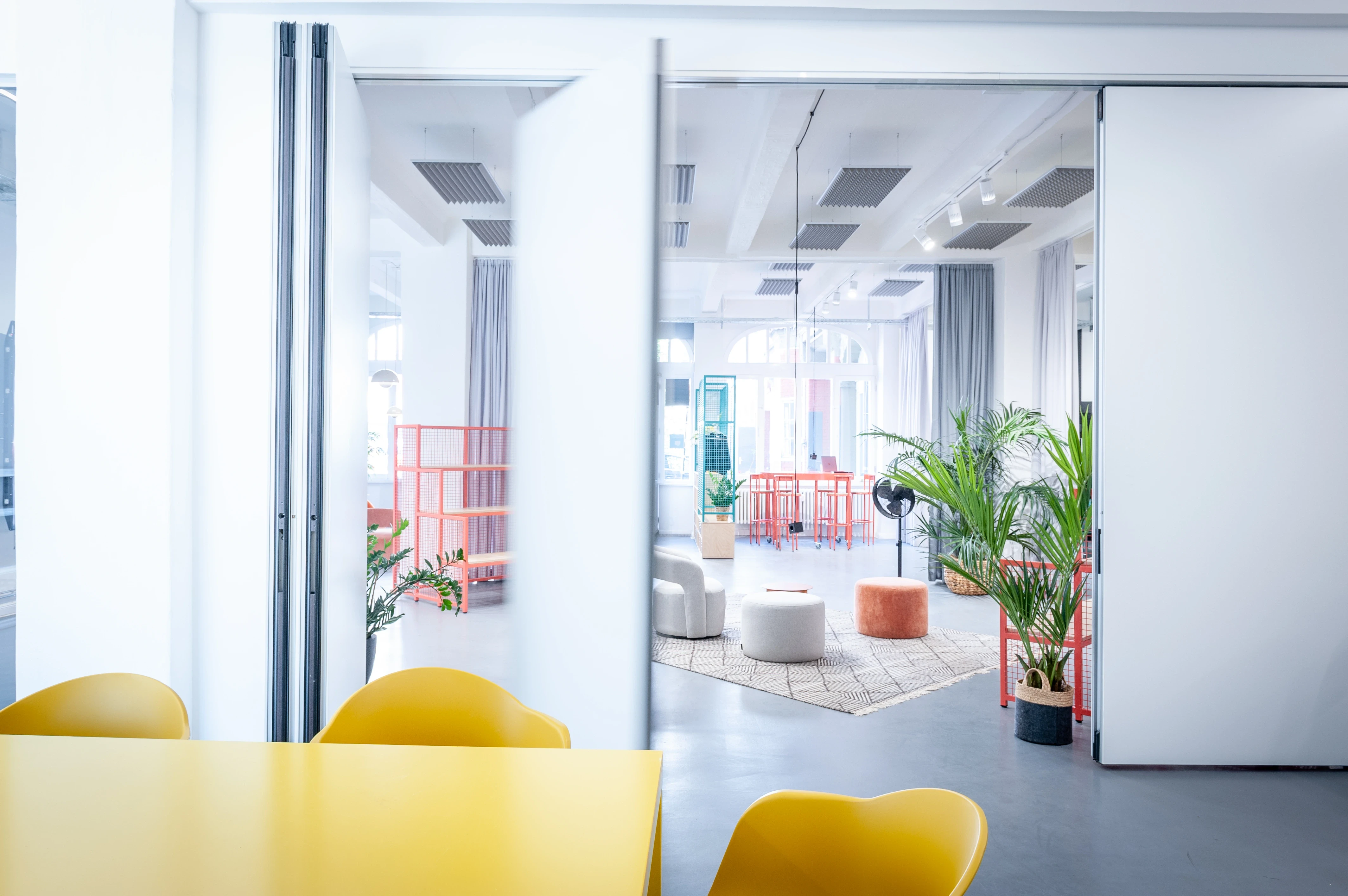 View from a yellow table through sliding glass walls into the open studio at HEY CONTENT STUDIO with lounge furniture, palm plants and coral wire shelves.