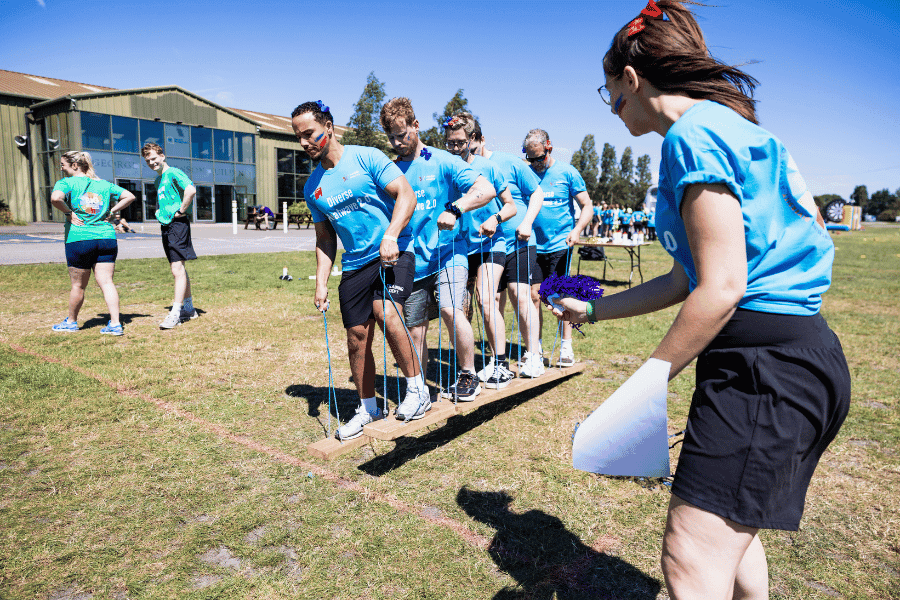 Outdoor team-building event with participants wearing matching blue shirts, taking part in inflatable obstacle courses and games, smiling and cheering, with the words 'ENTER A TEAM' in large bold white and green text across the image.