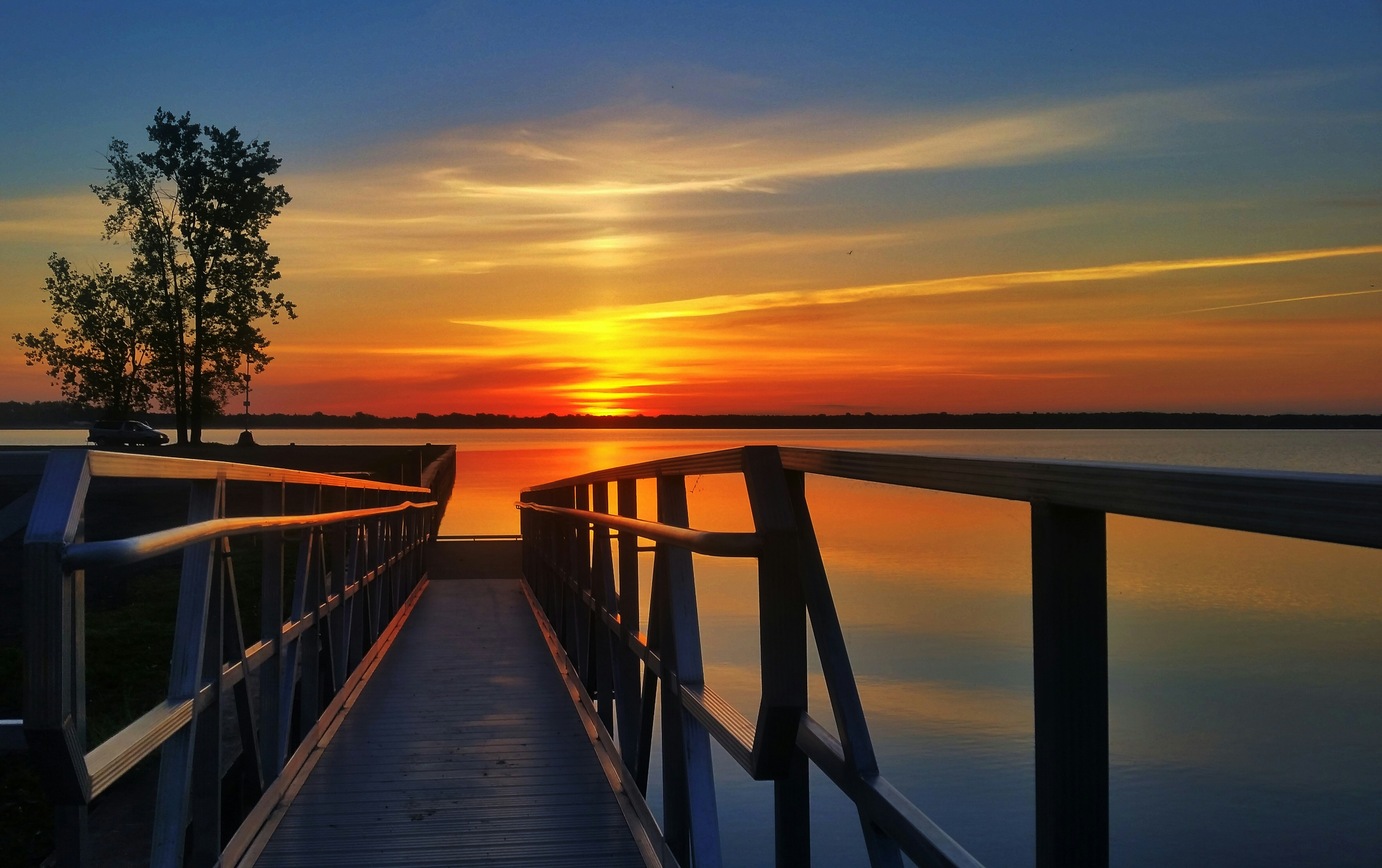A wooden pier leads out to a serene lake under a vibrant sunset, with orange and blue hues reflecting on the calm water, and a silhouetted tree standing to the side.