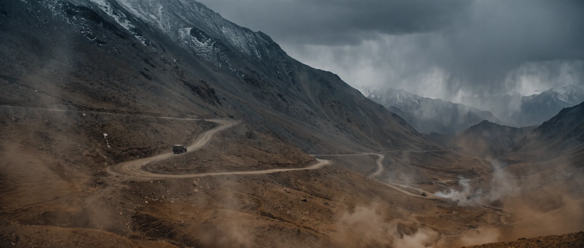 Concept art: a single vehicle on a switchback road cut into a bare mountainside under storm light.
