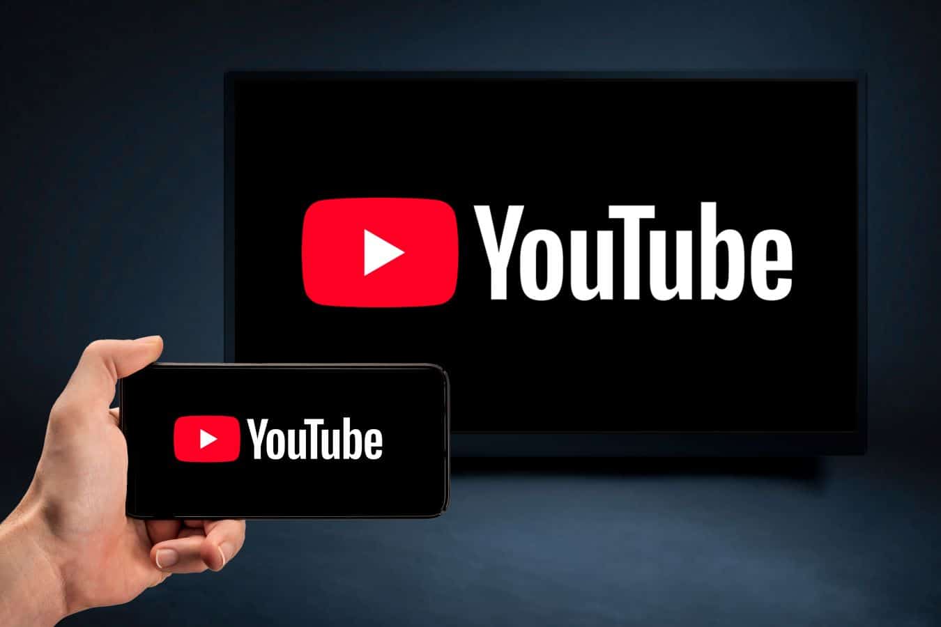 A hand holds a smartphone displaying the YouTube logo, with the same logo mirrored on a larger screen in the background, highlighting YouTube's multi-device compatibility.