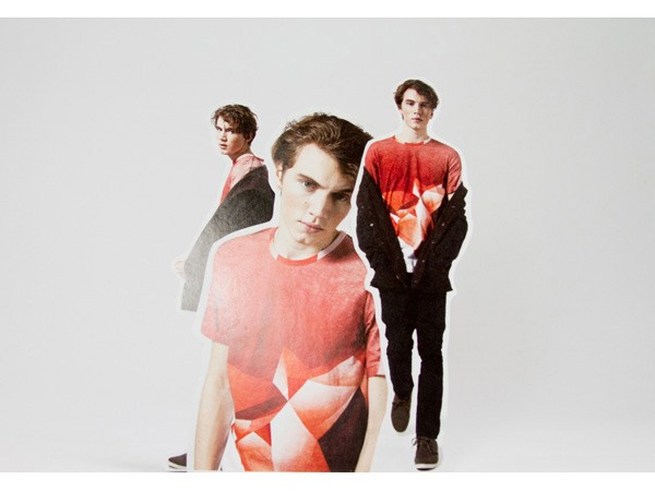 Three young men stand against a plain white background. The man in the foreground wears a red t-shirt with a geometric design. The man behind him wears a dark jacket over a white t-shirt. The man on the right wears a red t-shirt with a white geometric design and black pants. All three have short brown hair and are looking directly at the camera.