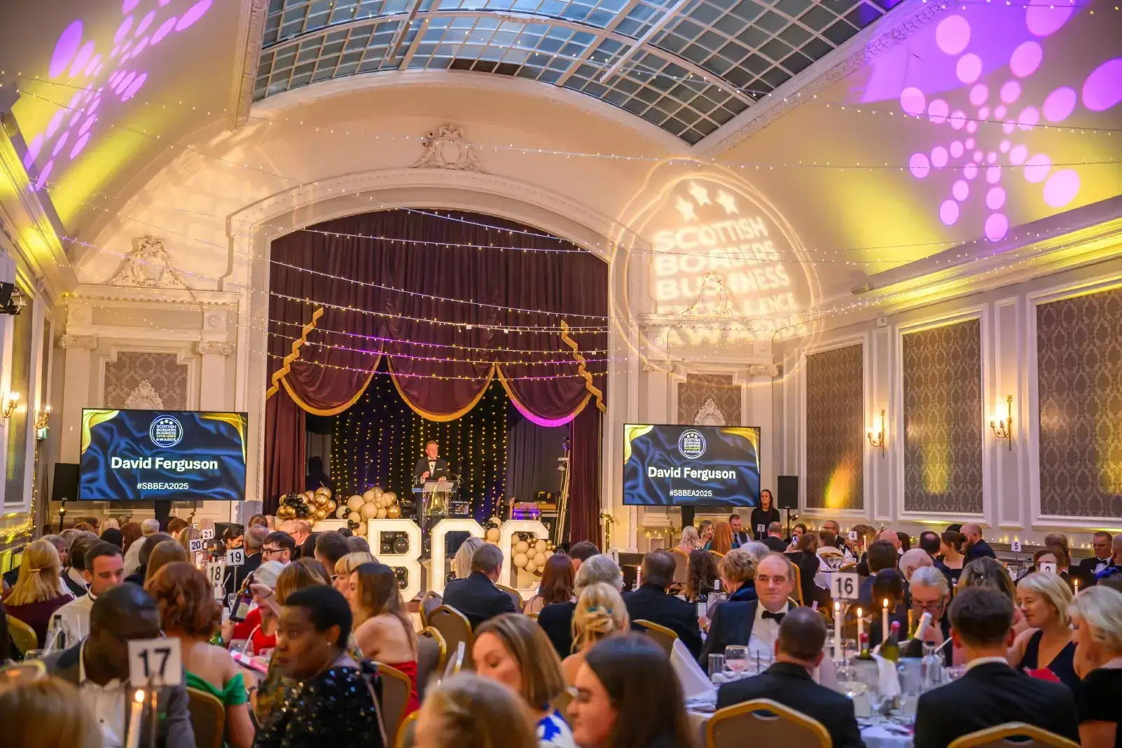 A grand ballroom is filled with elegantly dressed guests seated at tables for an awards ceremony. A stage with a podium is lit brightly, with a speaker presenting. The ambiance is festive, with decorative lighting and a projection reading "Scottish Borders Business Excellence Awards."