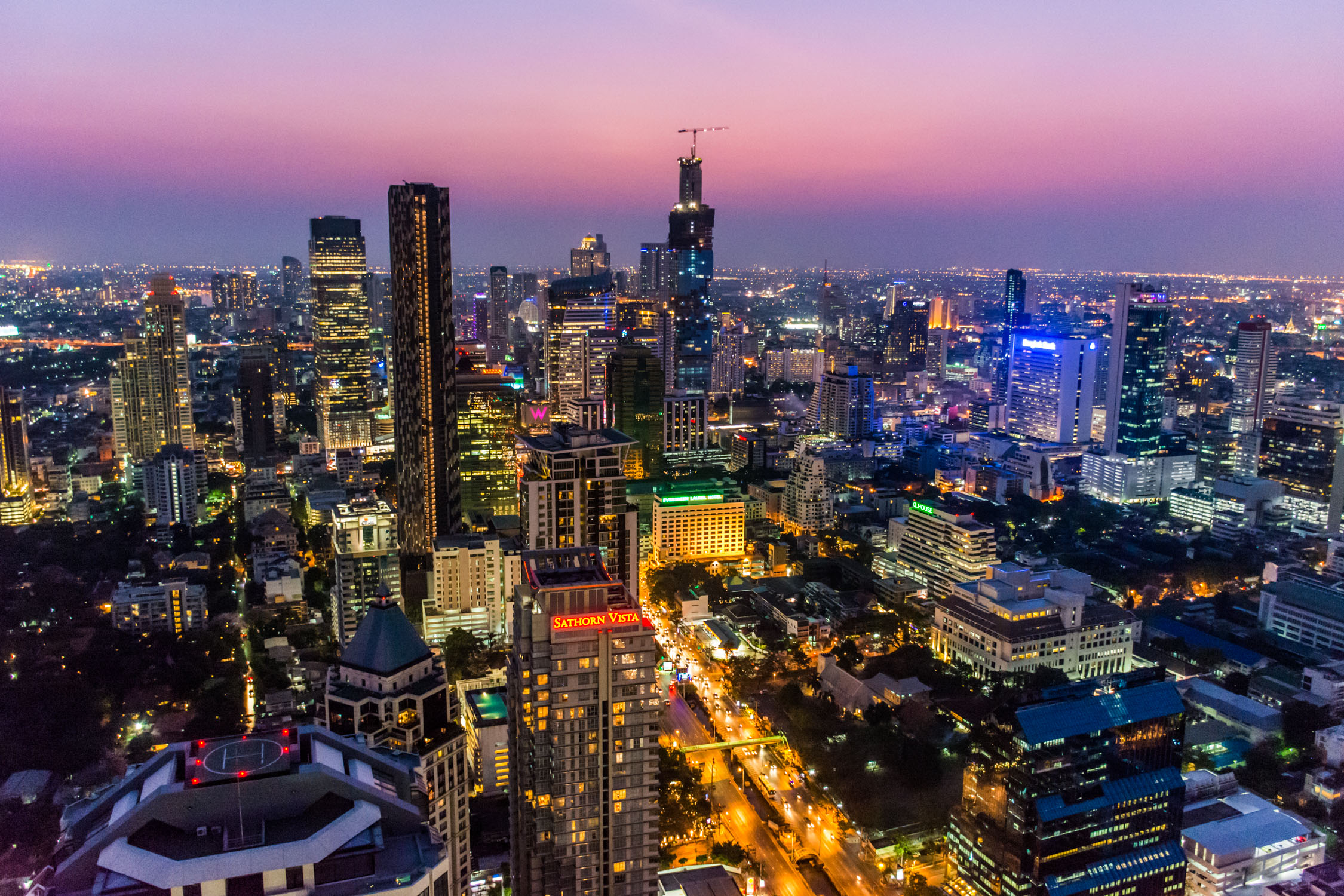 A wide cityscape at dusk with illuminated skyscrapers and a pink and purple sunset sky over a dense urban skyline.