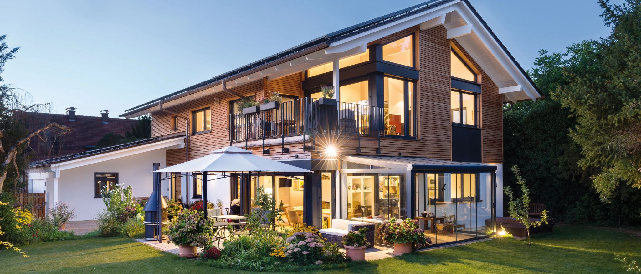 Modernes Holzhaus mit großem Glasanbau, Balkon und Gartenterrasse bei Dämmerung.