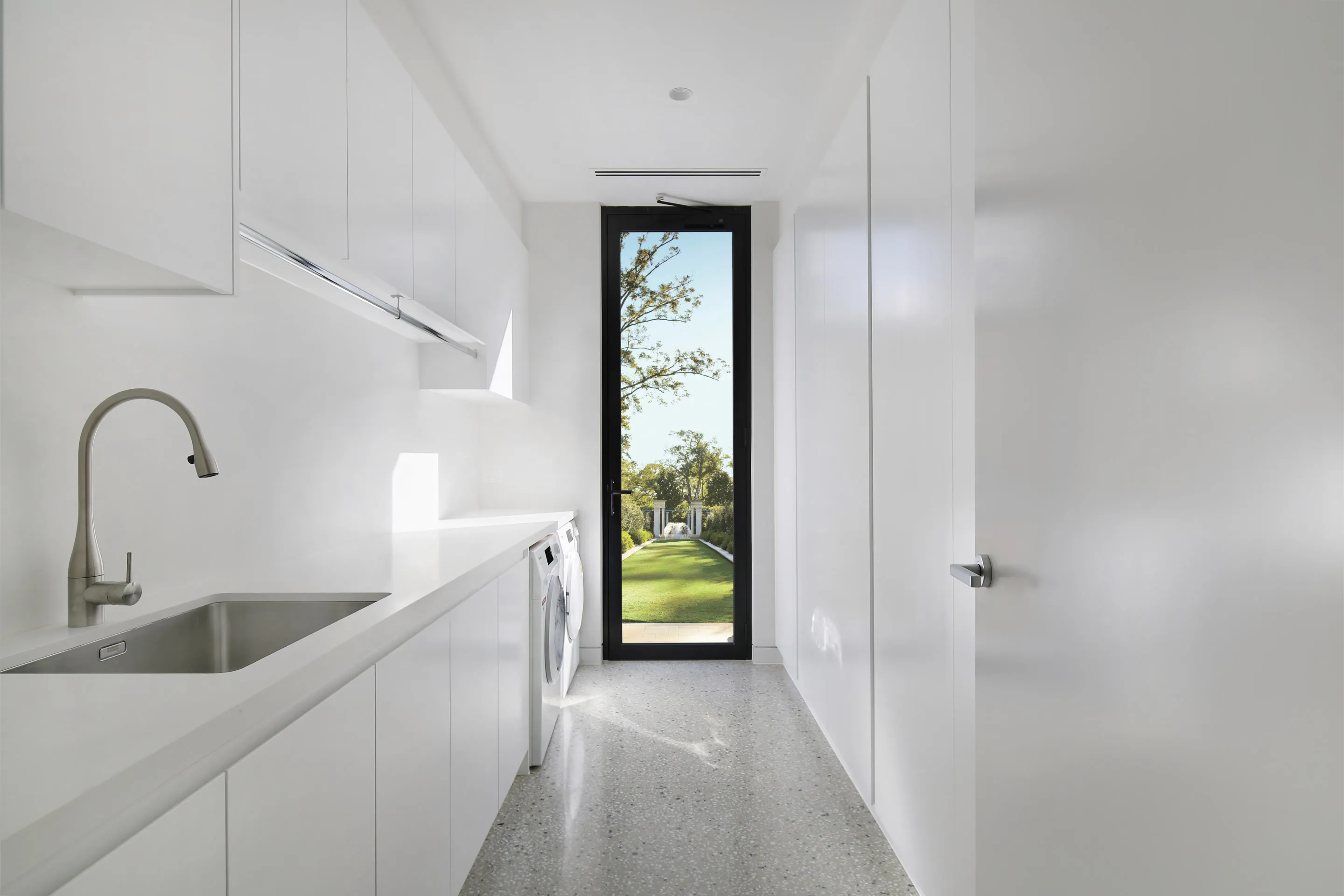 Galley laundry with white floor-to-ceiling cabinetry, a stainless sink and gooseneck tap, a stainless drying rail, terrazzo floor and a tall black-framed glazed door to the garden