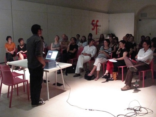 A photo of a group of students listening to a lecture given by a person standing in front of a laptop.