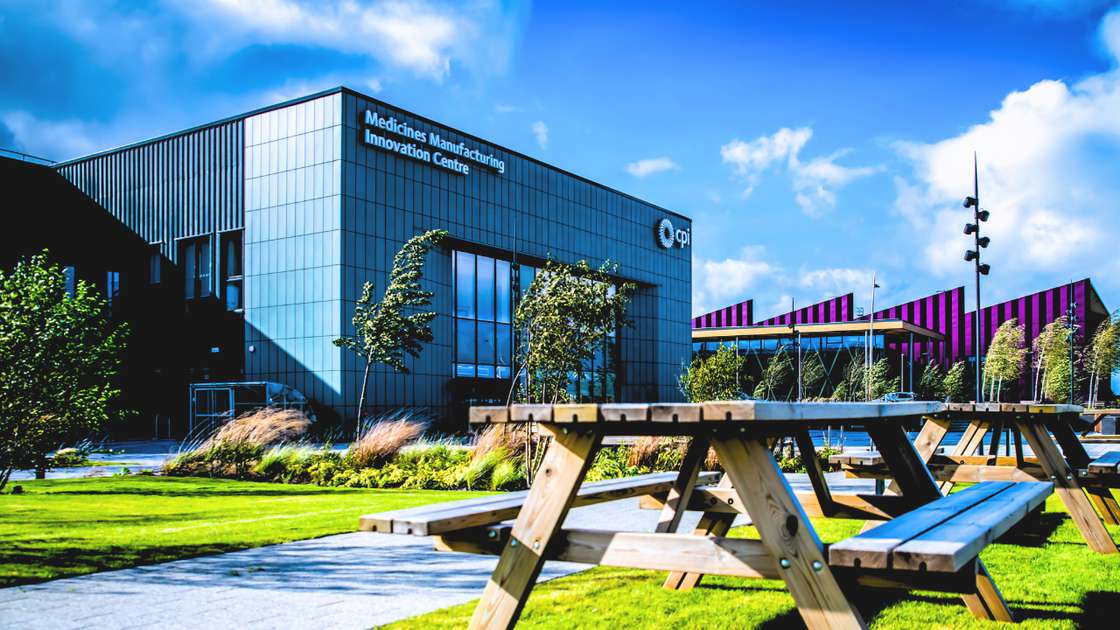 Exterior of CPI's Medicines Manufacturing Innovation Centre, a dark-clad building with wooden picnic benches on the grass under a blue sky