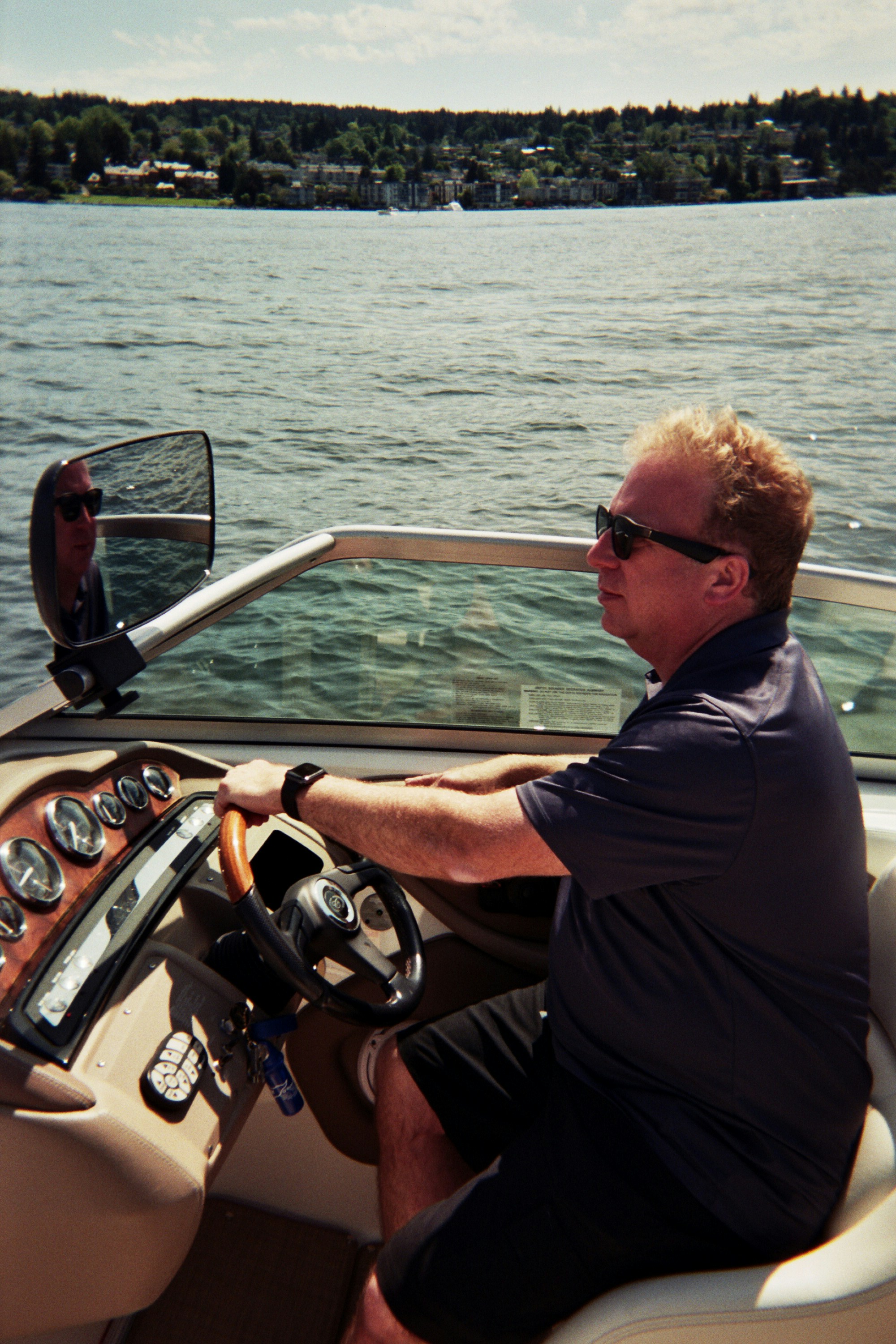 A person wearing sunglasses pilots a motorboat on a sunny day, surrounded by calm waters and distant leafy shorelines, highlighting a serene outdoor boating experience.
