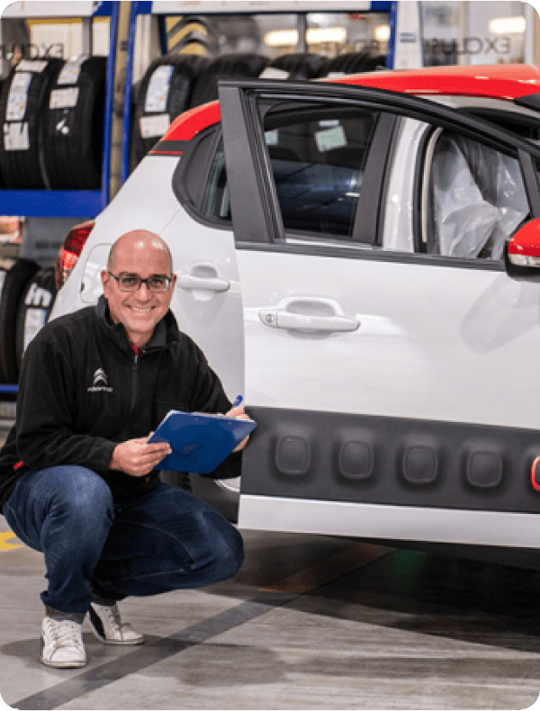 "Técnico de Citroën sonriendo mientras revisa un vehículo blanco con detalles rojos, en un taller con neumáticos al fondo."