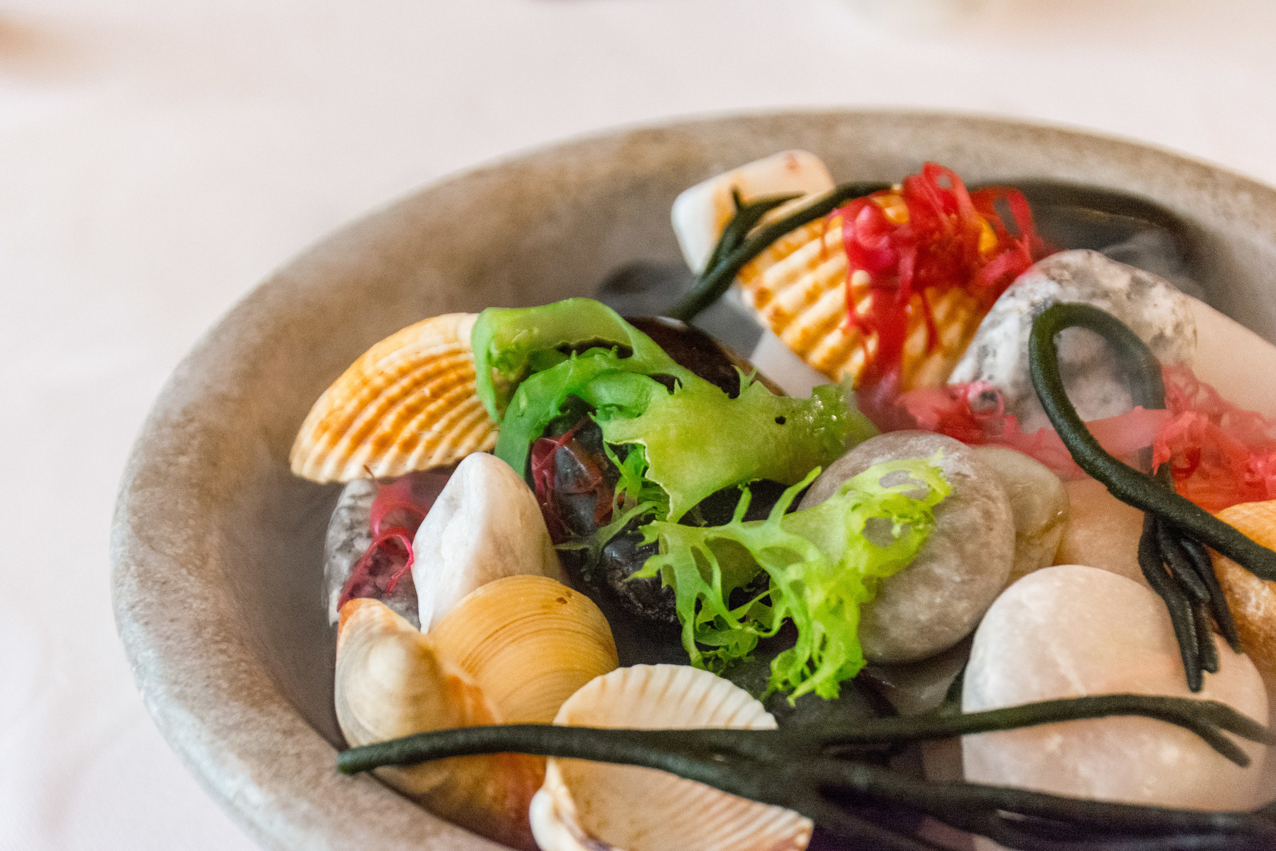 A stone bowl filled with steamed clams, green seaweed, and red chili peppers in a light broth.