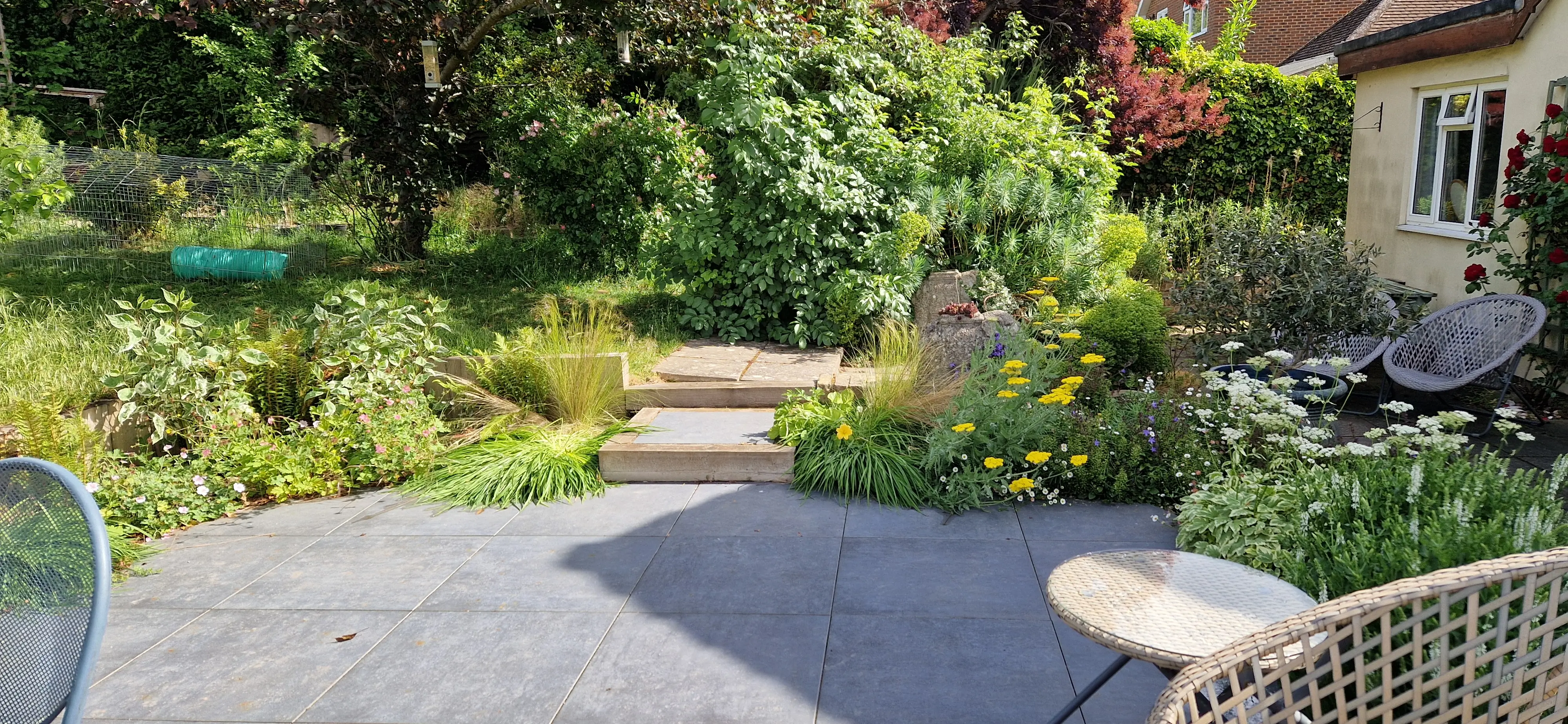 A scenic garden path surrounded by lush greenery and trees, leading to a cozy outdoor seating area.