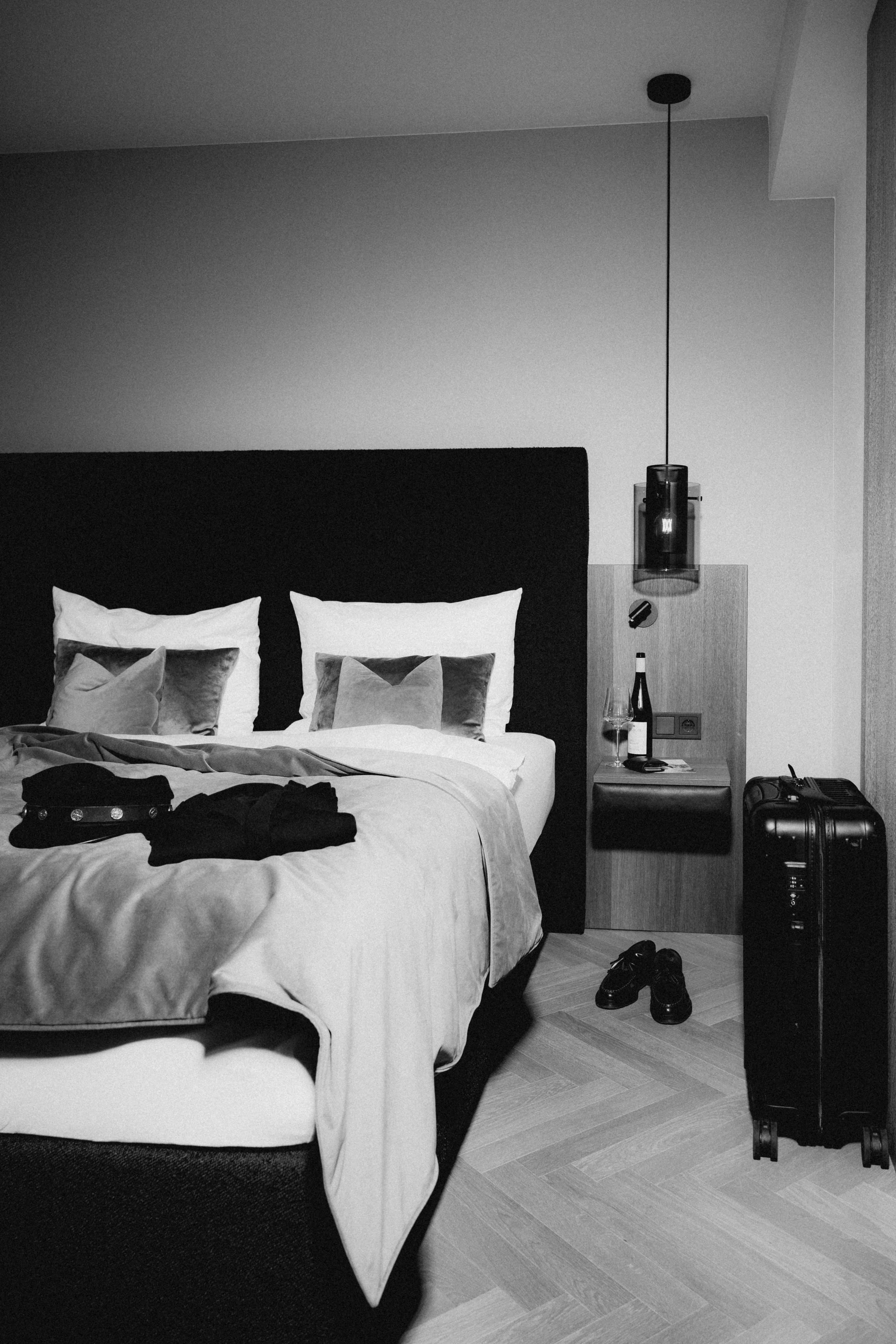 Schwarz-Weiß-Fotografie eines stilvollen Hotelzimmers mit gemachtem Bett, Gepäck und einer Weinflasche auf dem Nachttisch