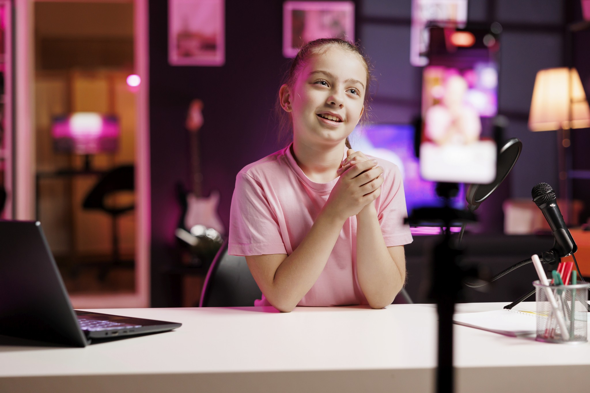 A child in a pink shirt sits at a desk with a laptop and microphone, speaking into a smartphone on a tripod in a room with ambient lighting and musical instruments.