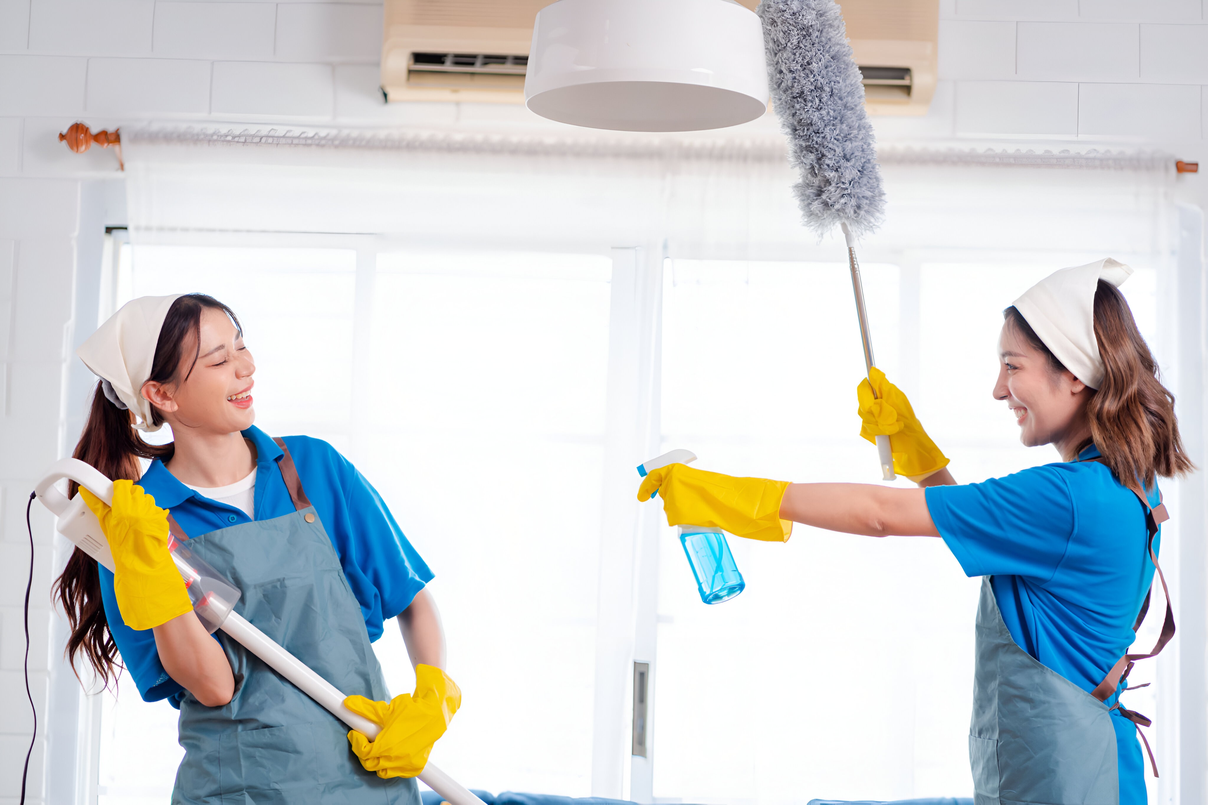 Two women in matching blue uniforms and yellow gloves playfully cleaning with a vacuum and duster, smiling while cleaning their home in a bright and airy space.