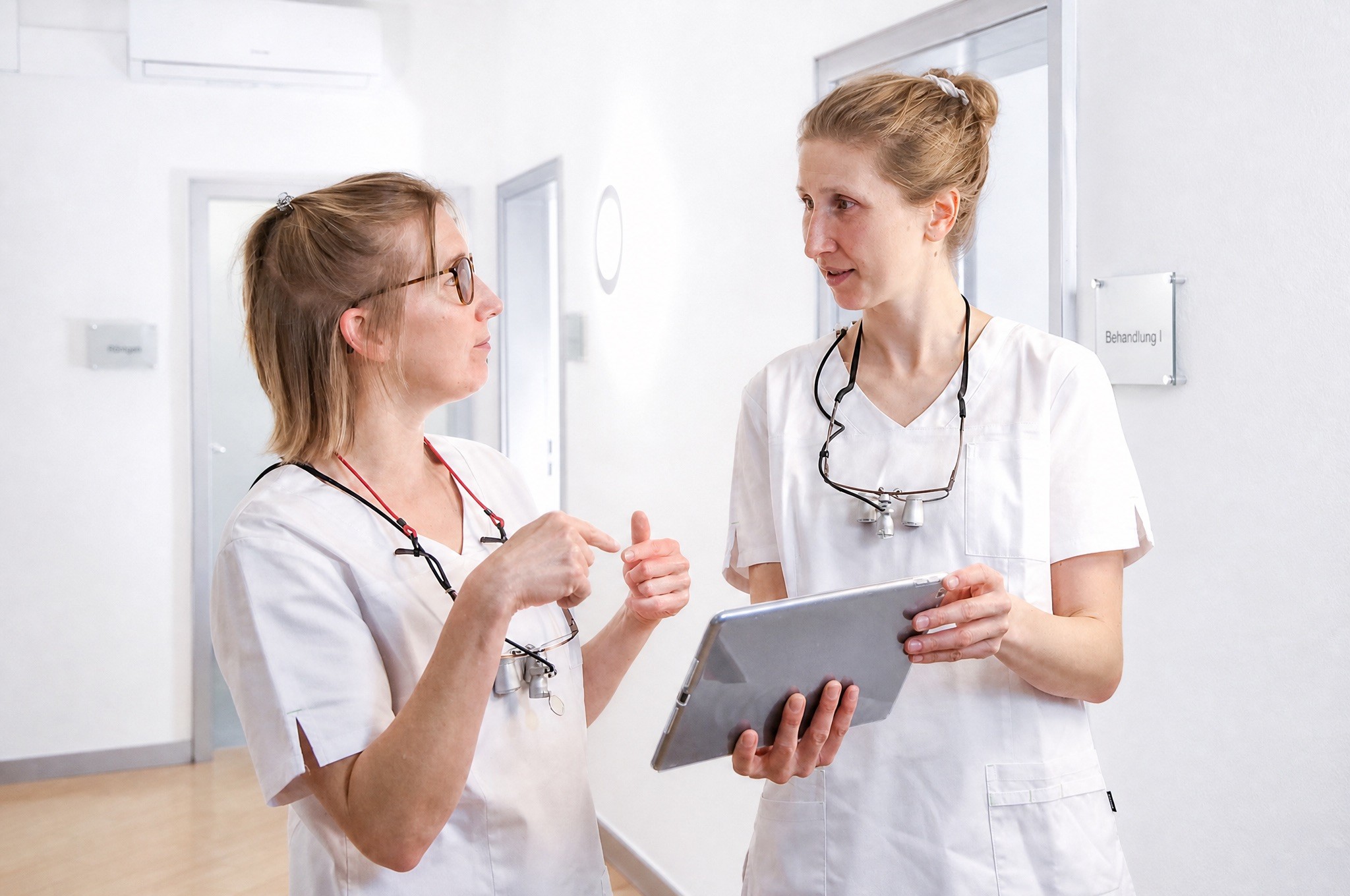 Two female dental professionals in white scrubs with dental loupes standing in a bright corridor, actively discussing details on a metallic grey tablet.