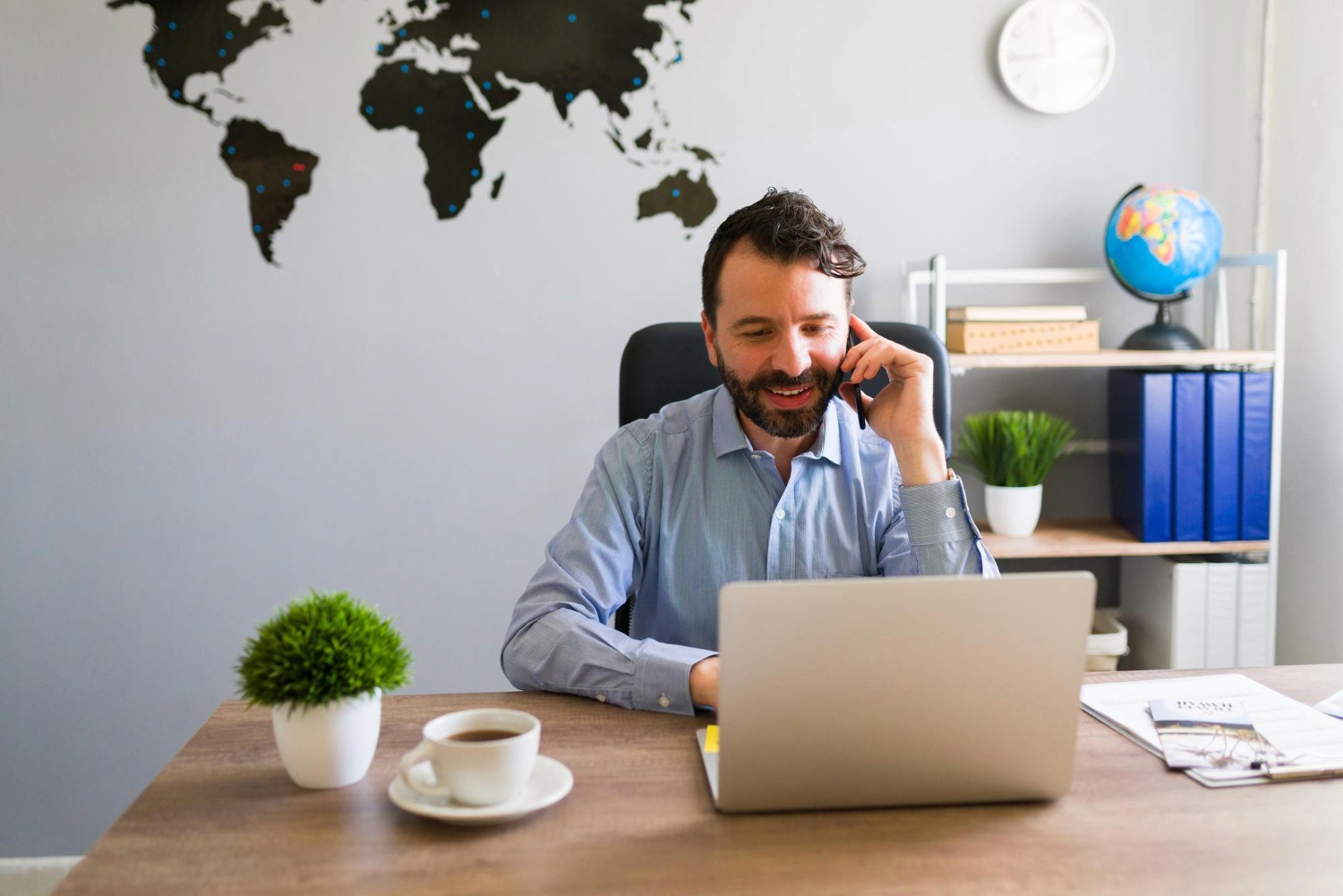 A man smiles while talking on the phone and using his laptop to complete his UAE visa cancellation from a modern office.