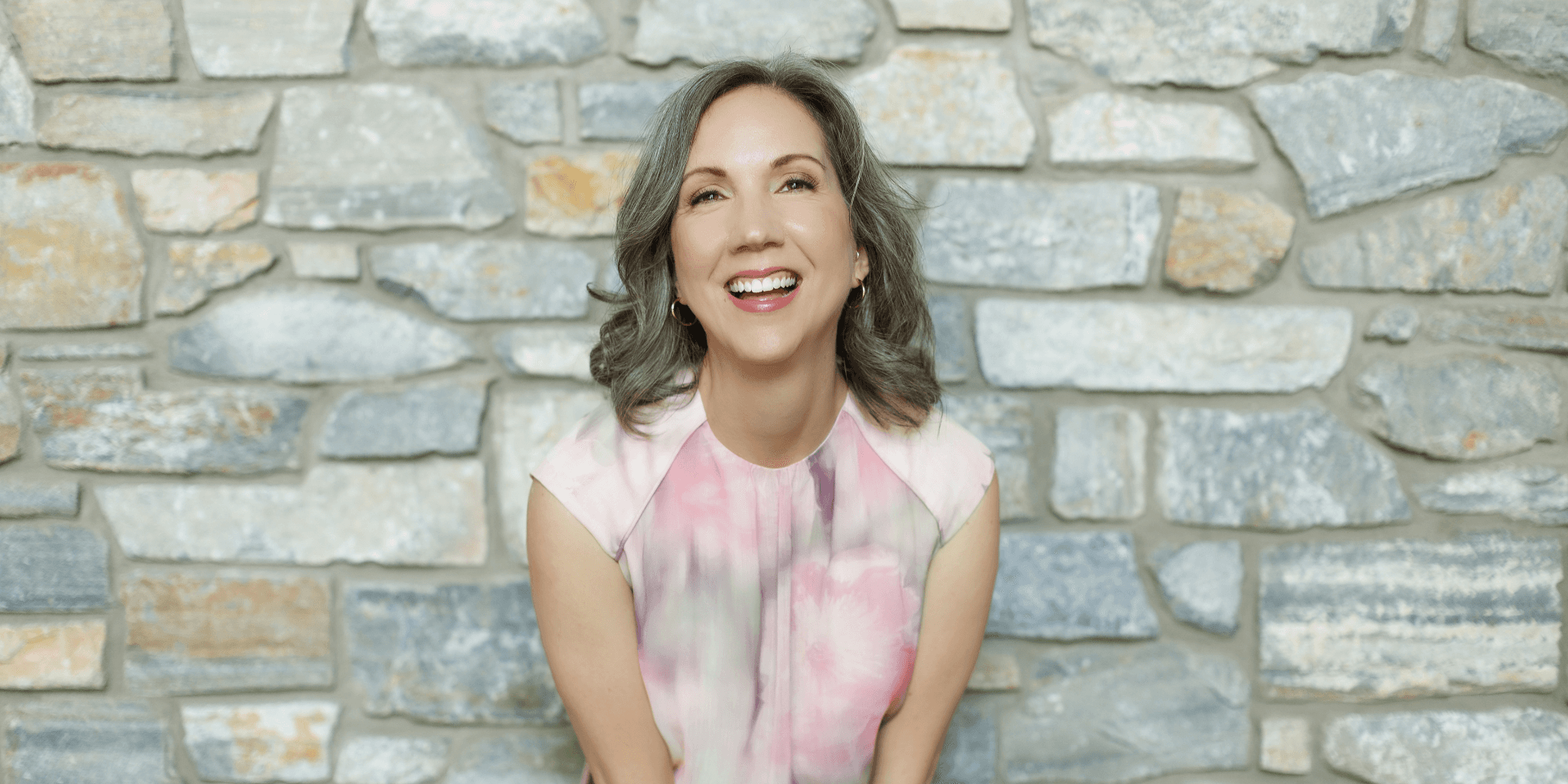 Smiling woman in pink blouse standing before a stone wall background, conveying warmth and confidence.