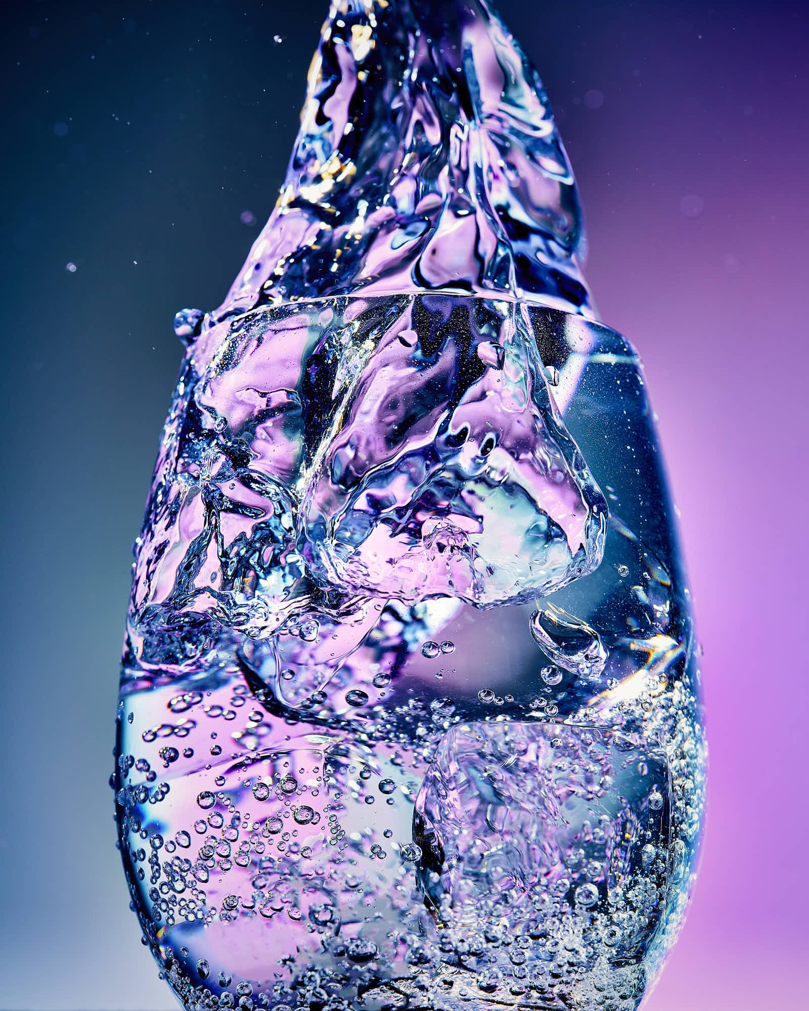 A colorful, translucent glass with ice and bubbles being dropped into it with vibrant purple and blue hues against a soft background.