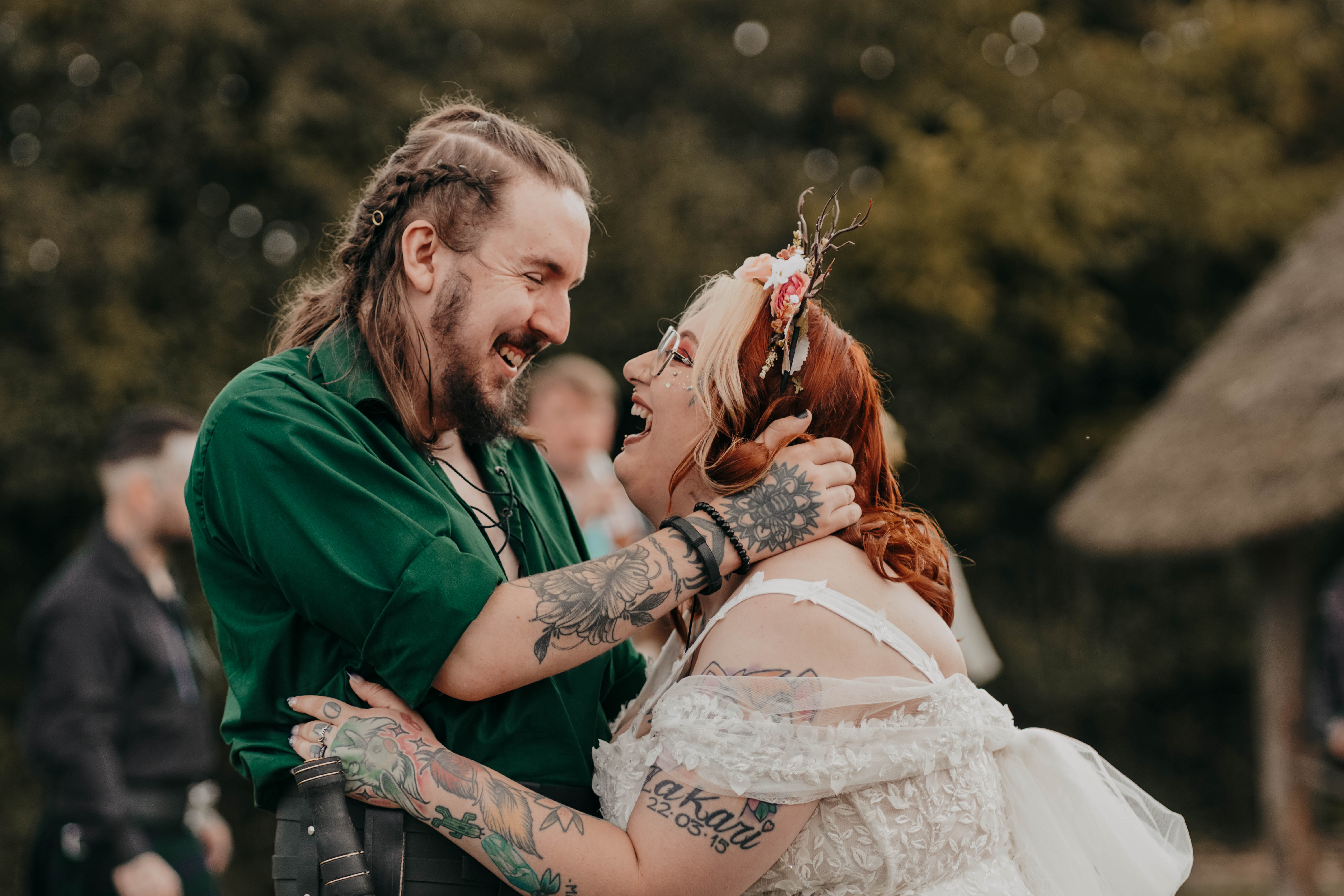 A candid, joyful photo of the bride and groom laughing together outdoors. The groom, in a green shirt, smiles down at the bride, who is looking up at him with a wide, happy laugh as they embrace.