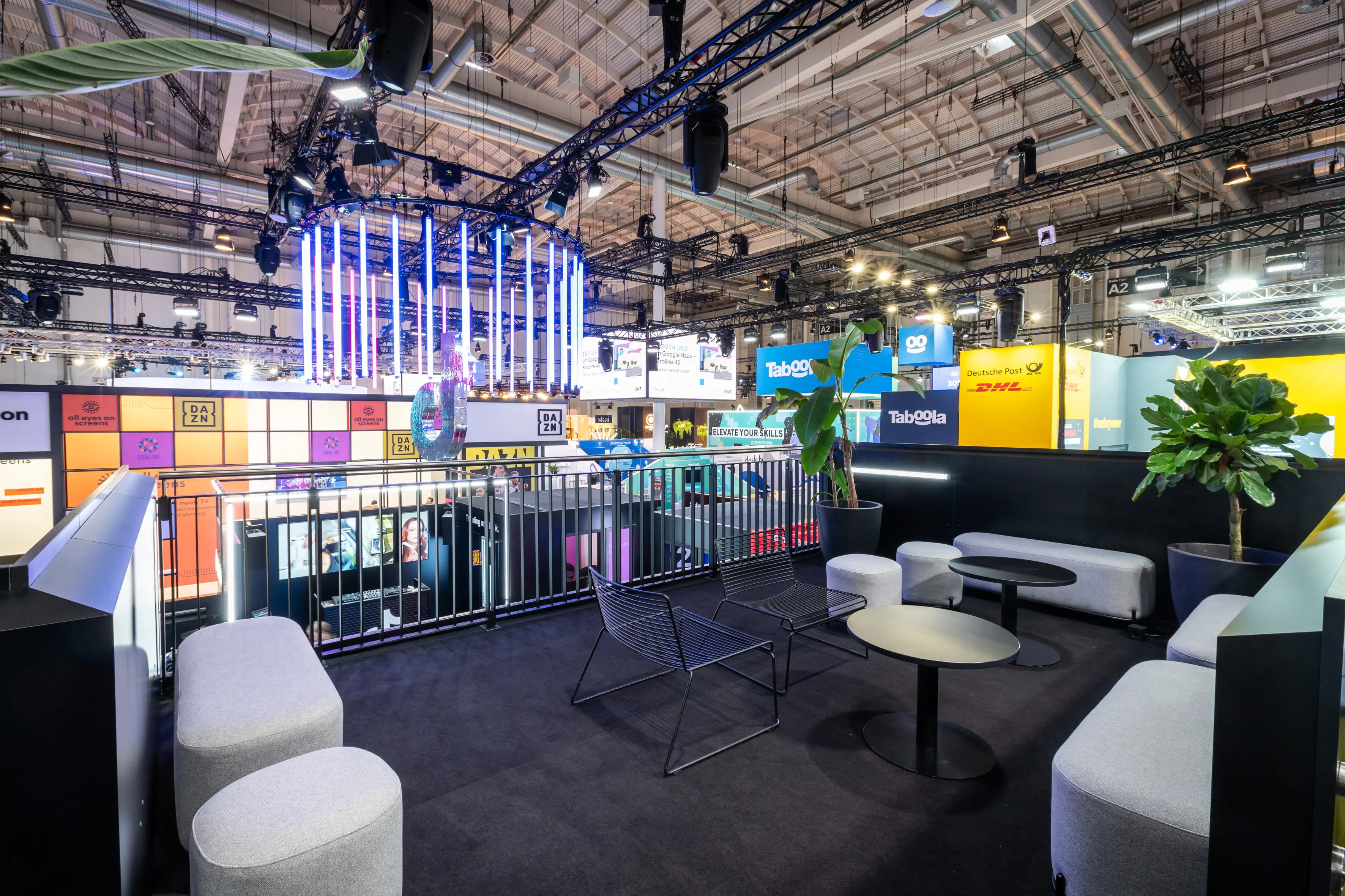 Upper-floor lounge of the TikTok booth with grey poufs, sofas and wire chairs overlooking the trade fair hall.