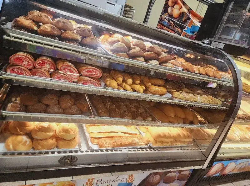 A variety of freshly baked goods, including pastries, donuts, and loaves, are displayed in a well-lit, glass bakery case, showcasing a tempting array of treats at Oneco Farmers Market, Bradenton, FL.