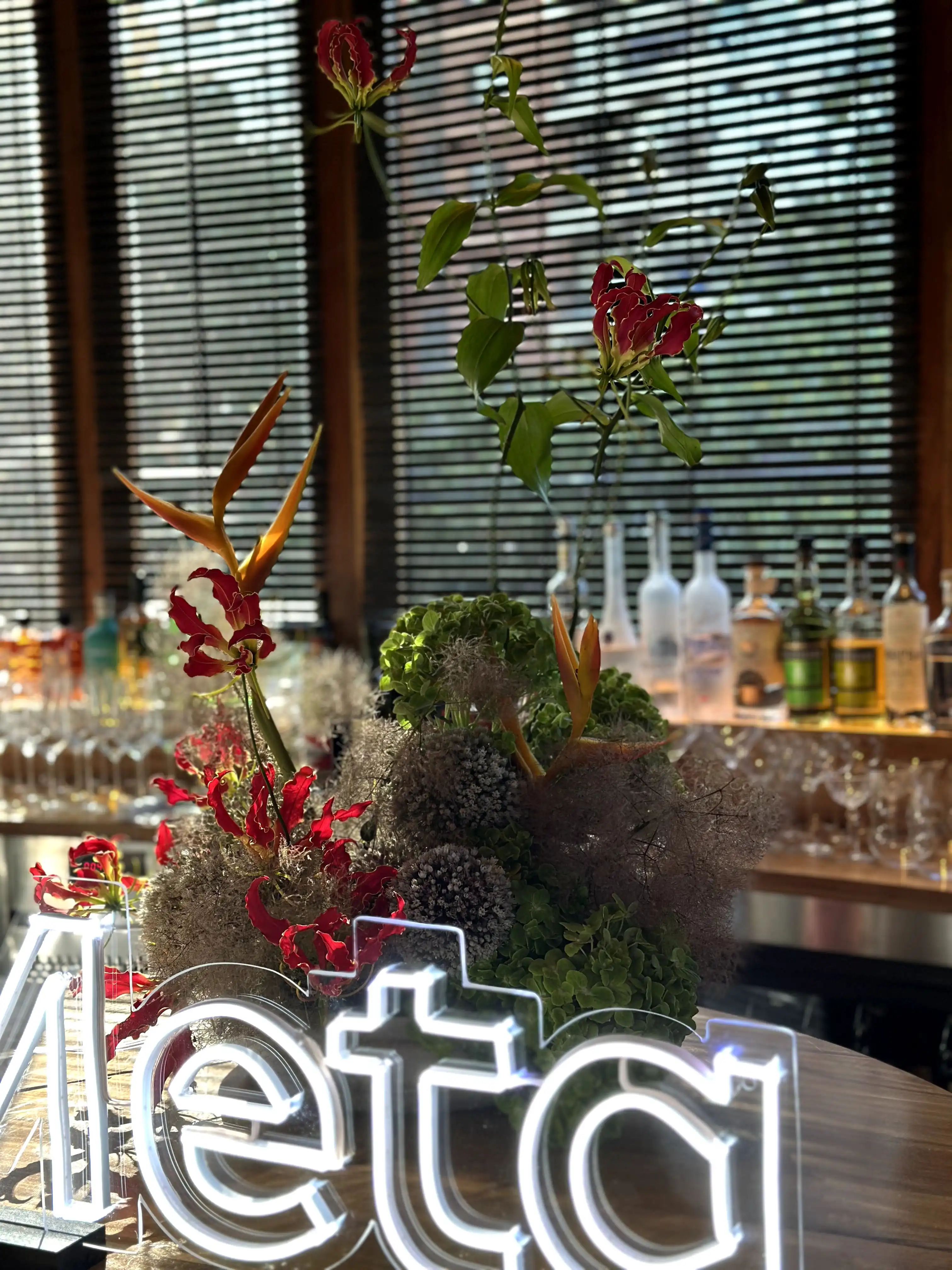Arreglo floral con gloriosas rojas, aves del paraíso y hortensias verdes sobre una barra de bar iluminada por un letrero de neón con la palabra ‘Meta’, al fondo botellas y copas tras una persiana de madera.