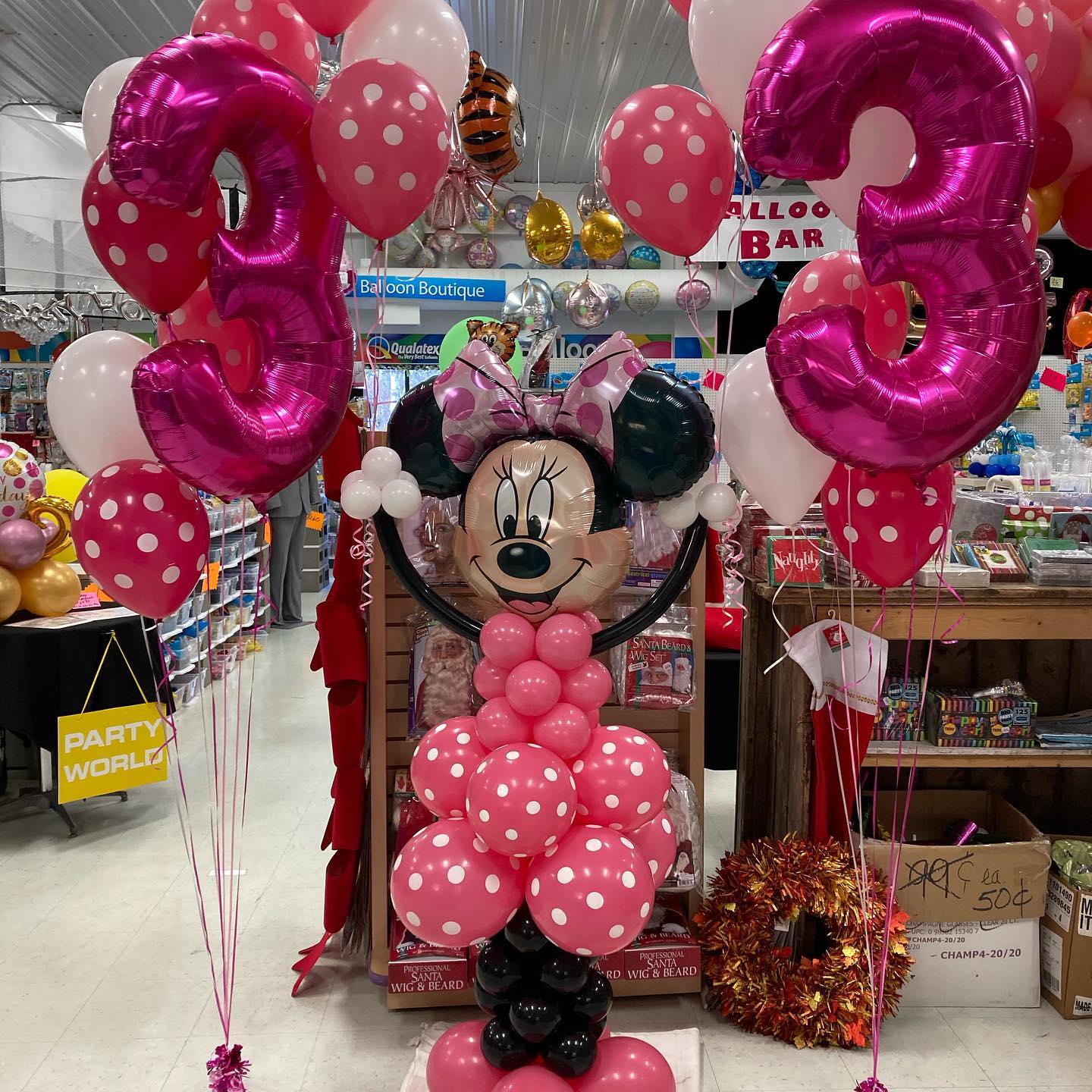 A colorful balloon display features a large number three in pink with polka dots, surrounded by themed character balloons and vibrant decorations, set in a festive party supply store.