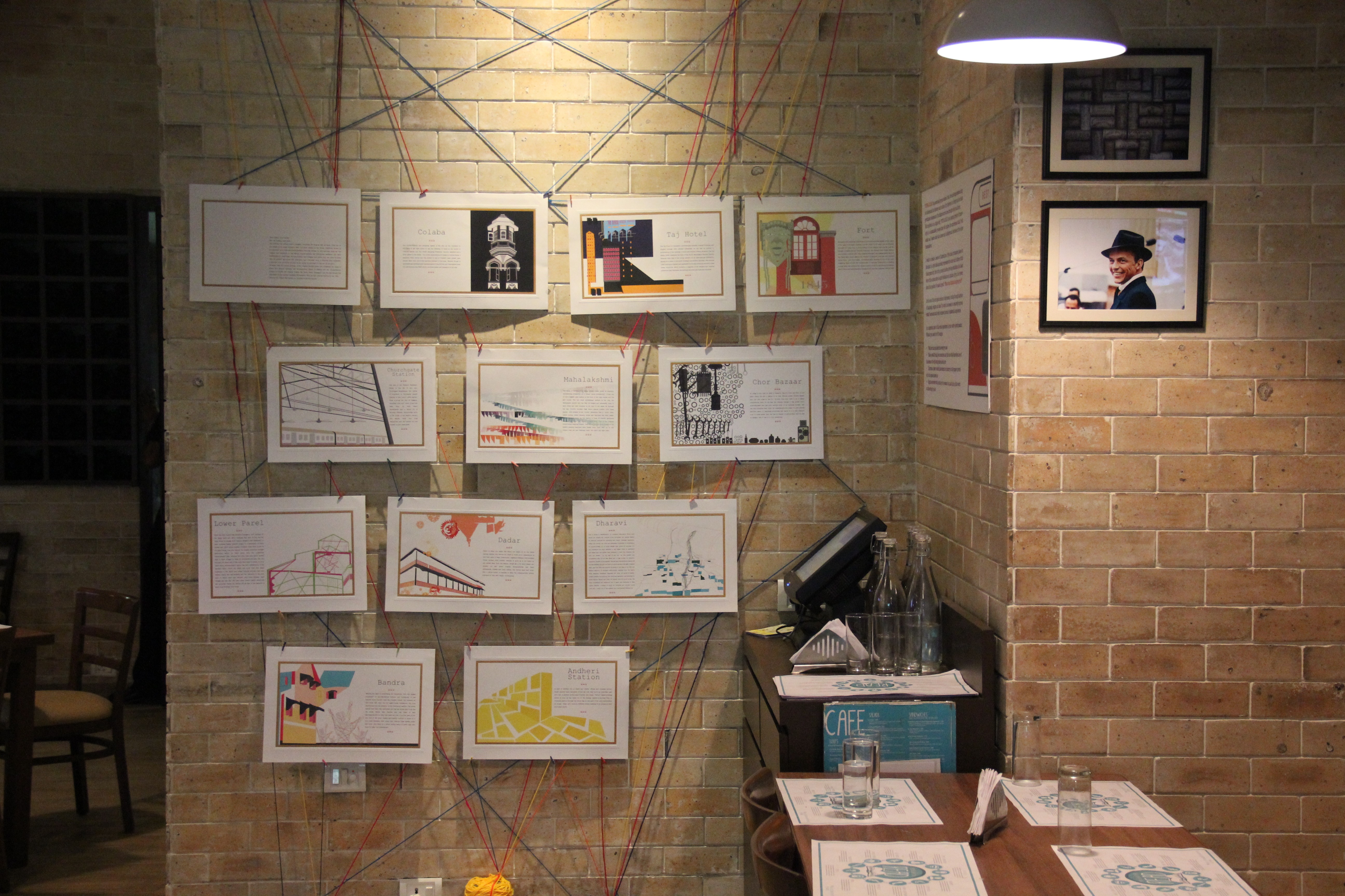 Different paper drawings suspended by wires on an illuminated brick wall.