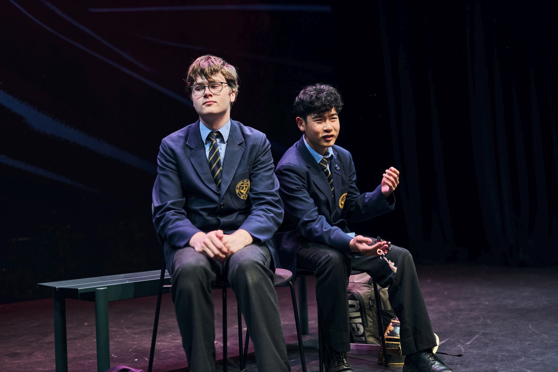 Two young actors wearing school blazers sit on a bench mid-scene. One sits stiffly with a neutral expression, while the other speaks animatedly, suggesting a contrasting character dynamic during a school-based performance.