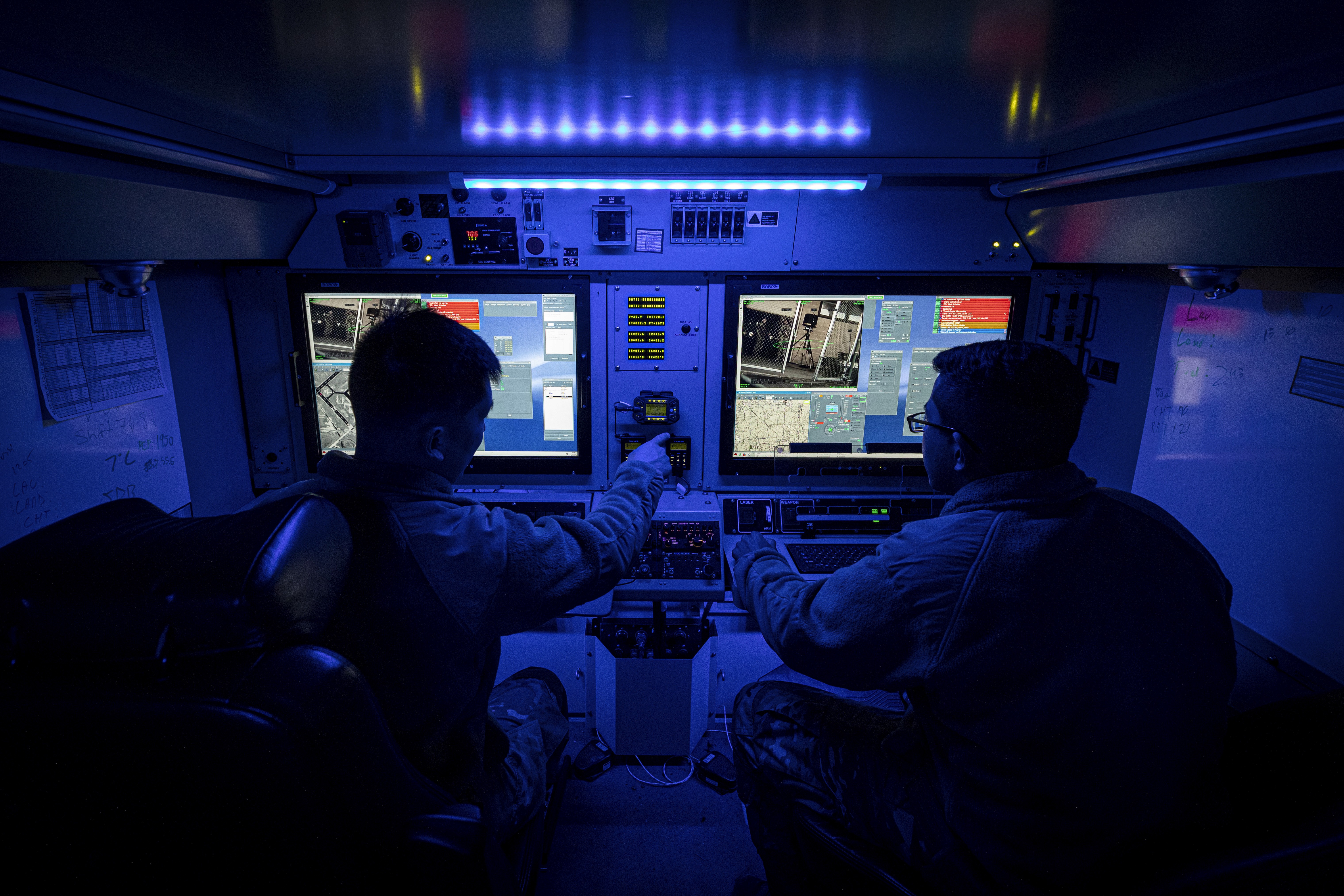 Two National Guardsmen sit next to  each other in a ground control station with mission controls screens in front of each