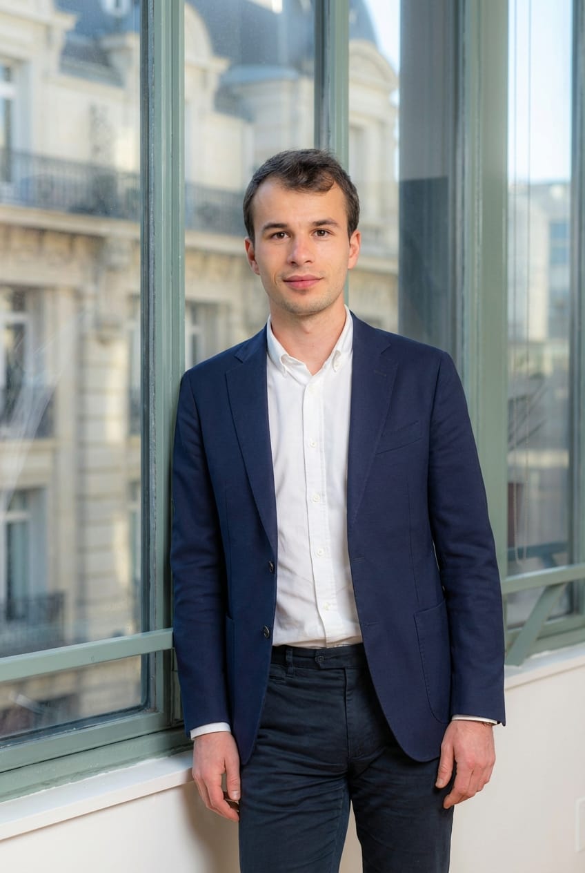 Young person with curly hair wearing a rust-colored blazer and black turtleneck, looking confidently at the camera against a neutral background.