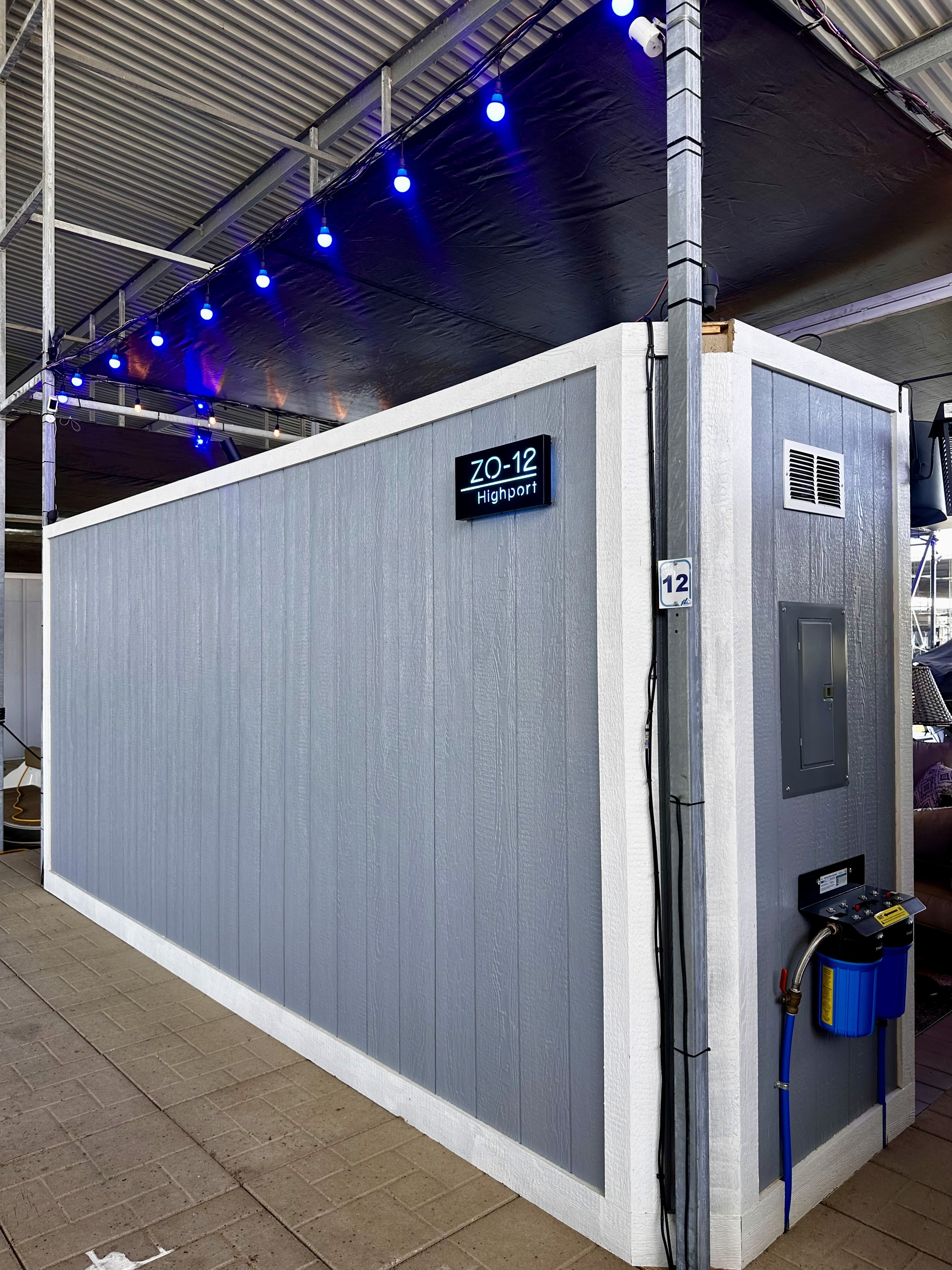 A sleek gray shipping container is repurposed into a functional space, adorned with vibrant blue lights overhead, situated in an industrial setting with exposed metal beams and a numbered sign indicating ZO-12 Highport.