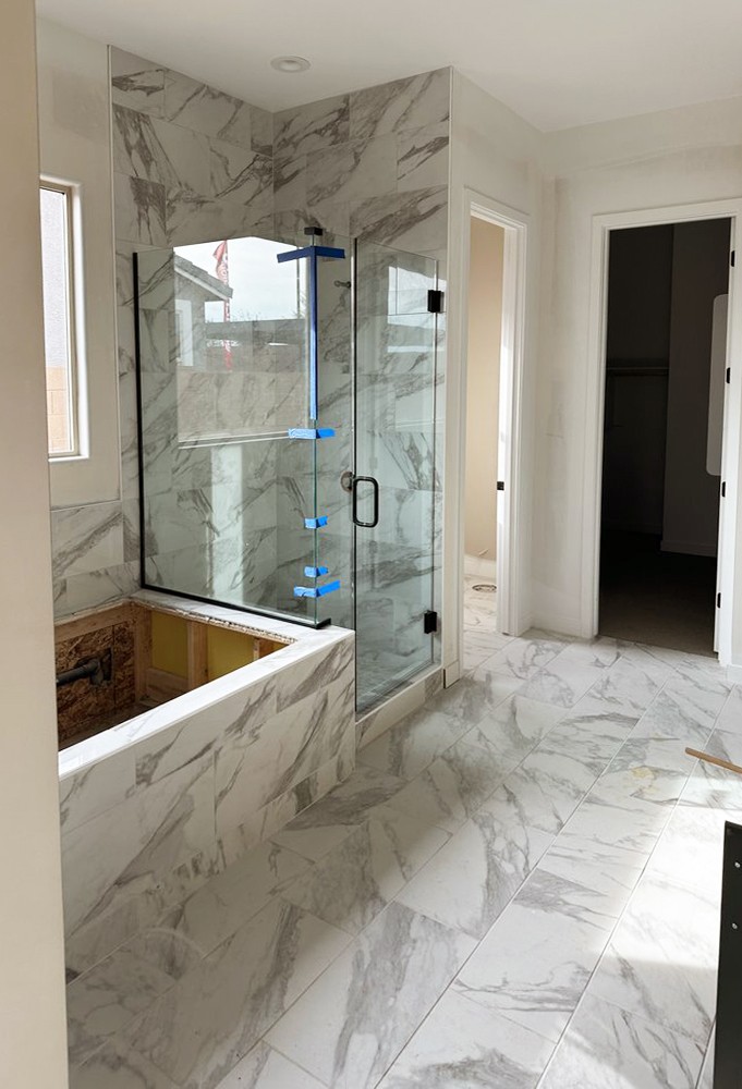 Luxurious bathroom with full marble-look tile flooring and walls, frameless glass shower enclosure, and a partially finished bathtub surround in matching tile.
