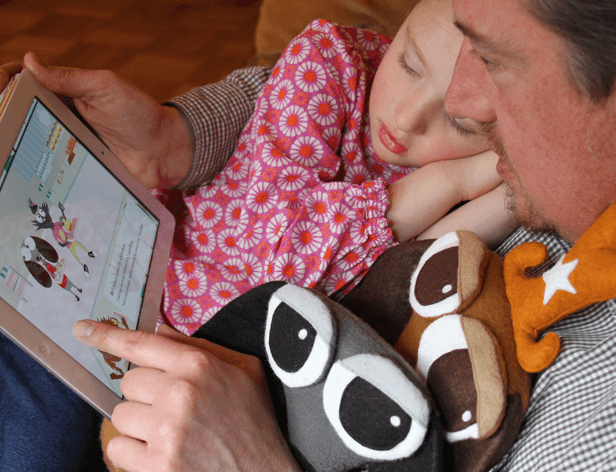 A man holds a tablet in his left hand, displaying a cartoon image. A baby, wearing a pink floral dress, rests on his lap, eyes closed. The man wears a checkered shirt and holds a stuffed animal with an orange star on its back. The setting appears to be a wooden floor.