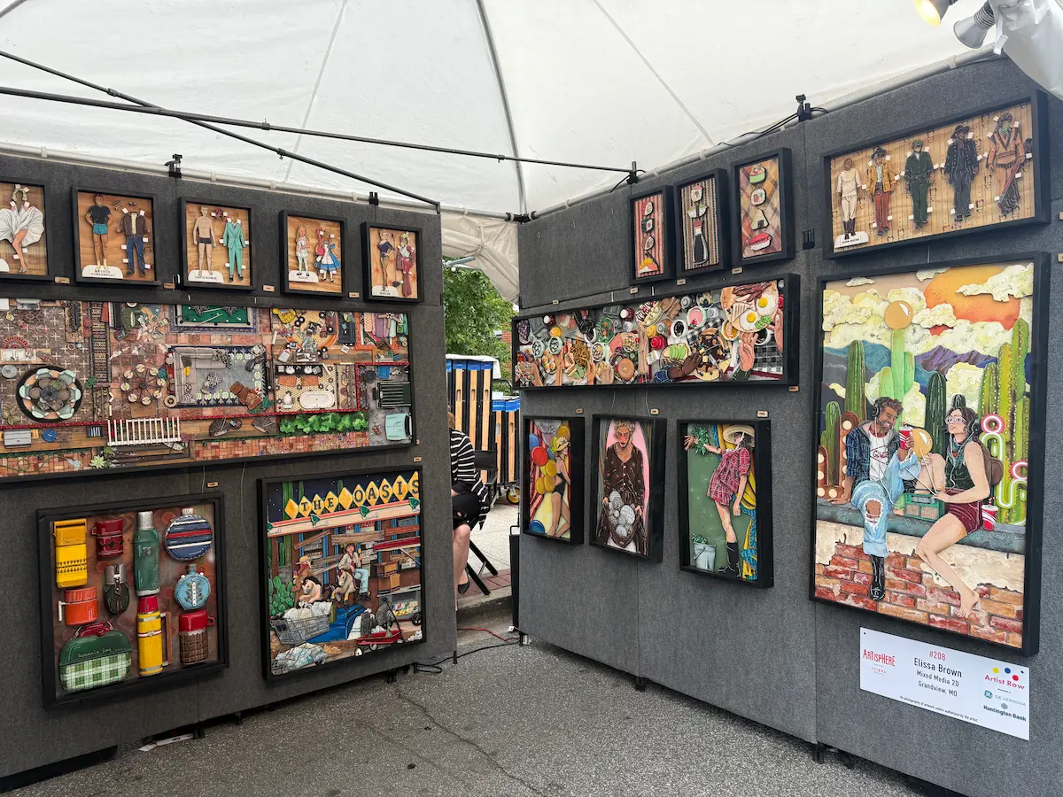 Mixed-media artwork displayed inside an artist booth at Artisphere in downtown Greenville, featuring detailed collage-style scenes, colorful objects, and handcrafted shadowbox pieces.