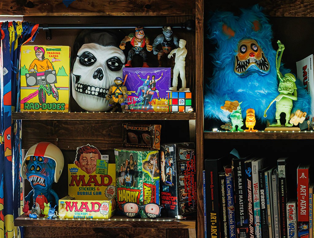 Wooden shelf filled with colorful collectibles, including a blue fuzzy monster figure, a Misfits skull mask, Rad Dudes trading cards, MAD magazine boxes, and vintage toys surrounded by art and design books.
