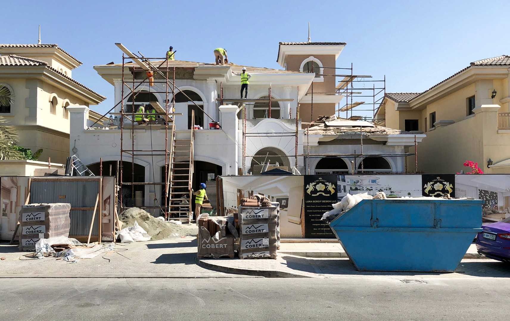 A construction site with workers in safety gear is actively renovating a large, two-story house with scaffolding and ladders against its facade, surrounded by building materials and a blue dumpster in the foreground.