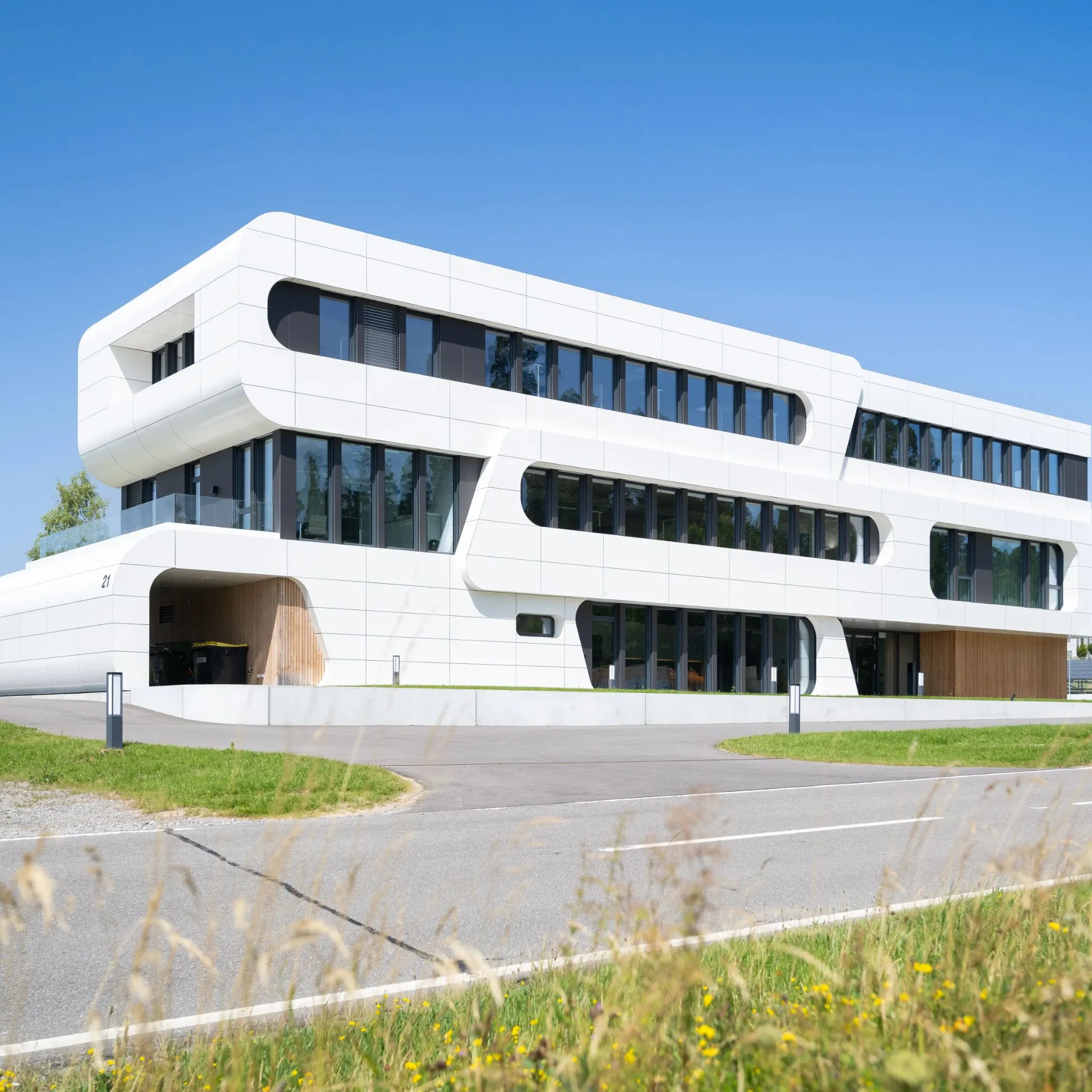 Modernes weißes Bürogebäude mit geschwungener Fassade und großen Fensterfronten unter blauem Himmel.