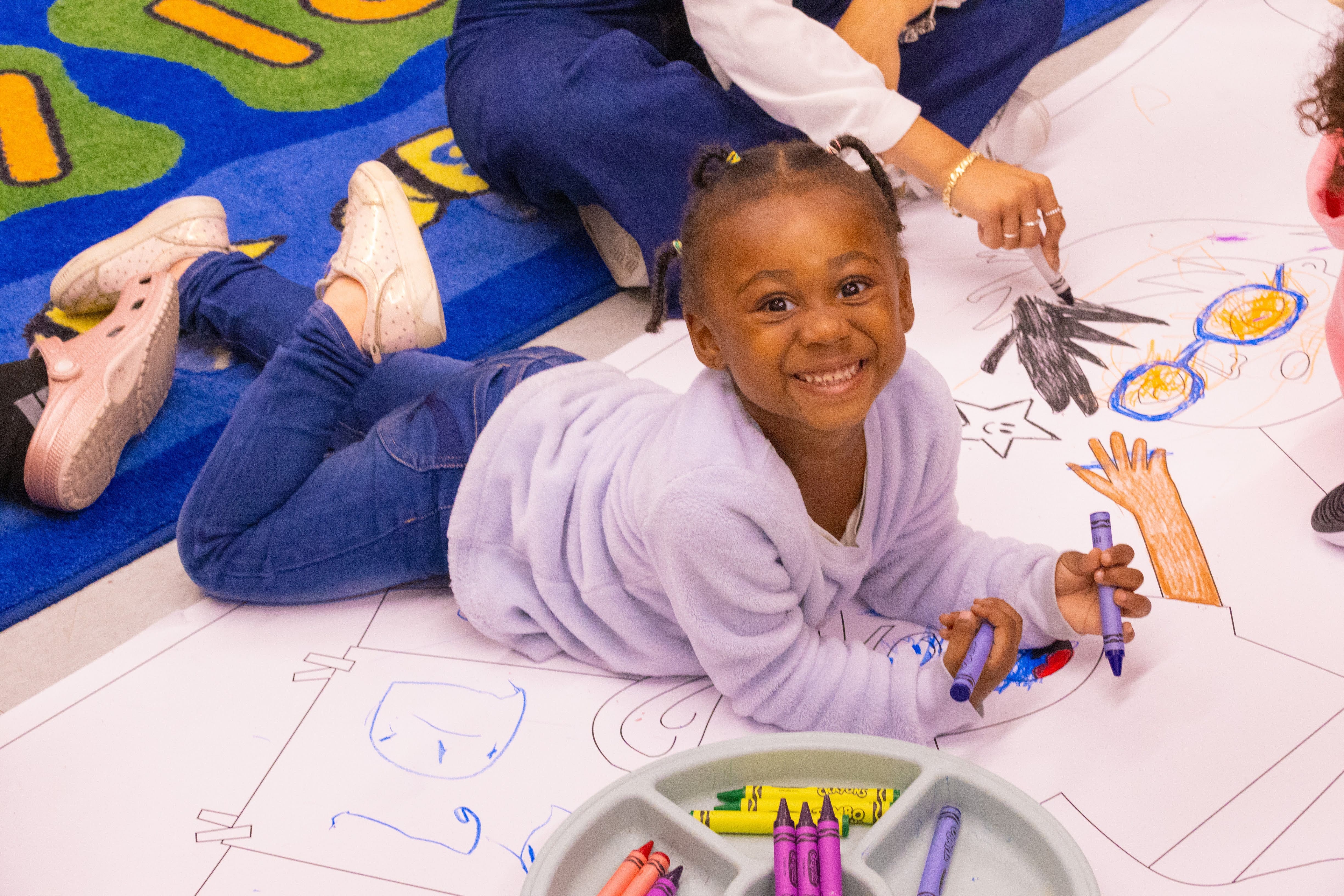 A little girl laying down holding crayons and coloring on a sheet of paper