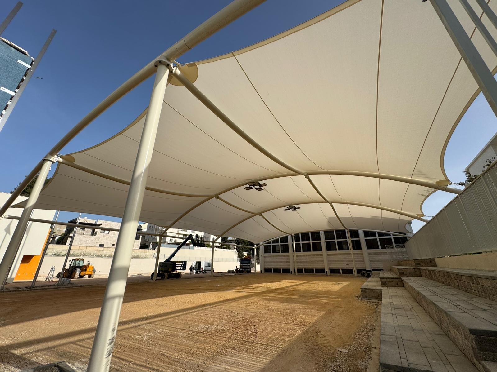 Large beige tensioned sunshades over an open sandy area with surrounding steps and buildings in the background.