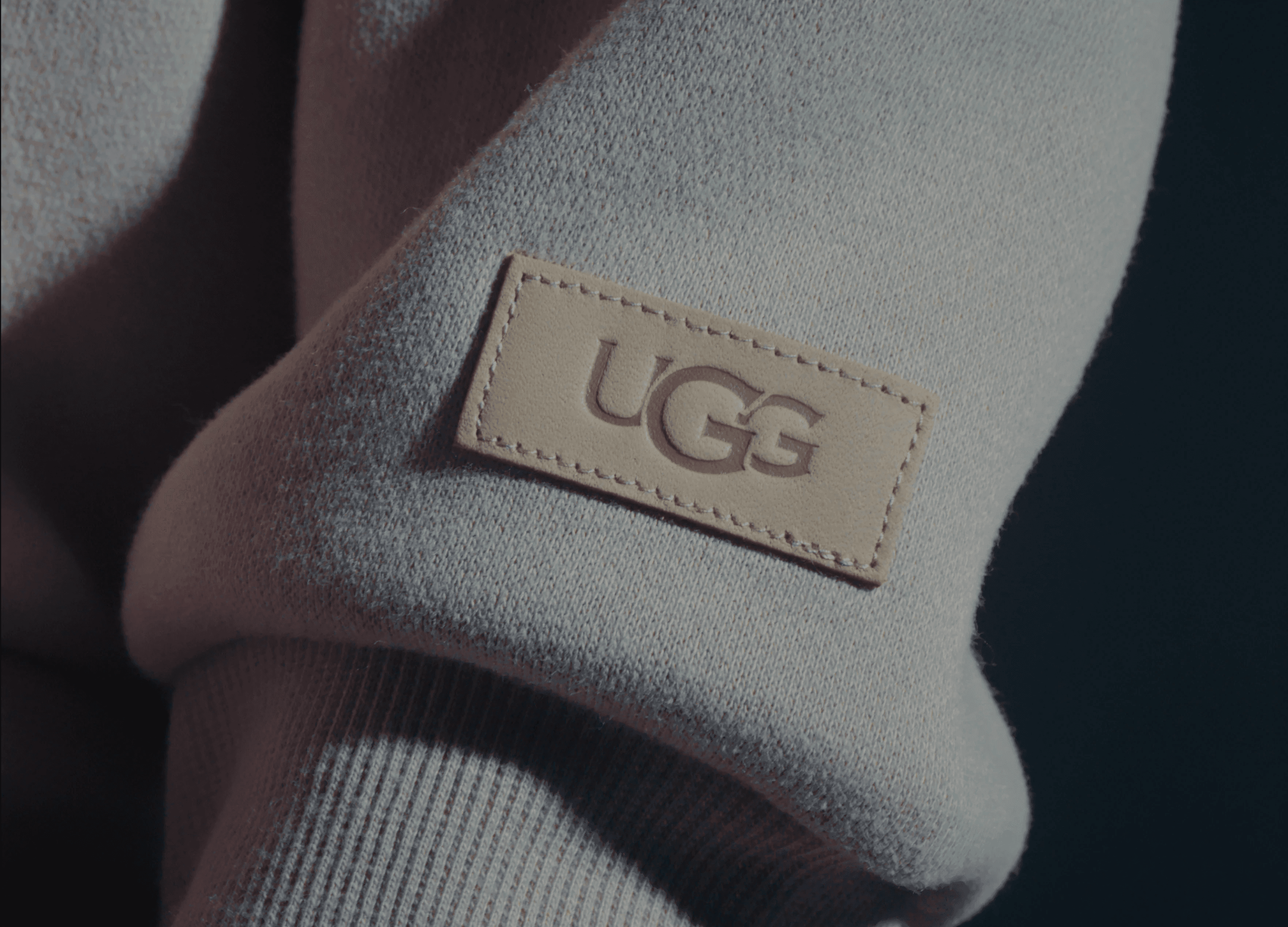 The image shows a close-up of a cozy, light-colored fabric with a stitched leather label featuring the UGG logo, highlighting the texture and softness for a luxurious, comfortable feel.